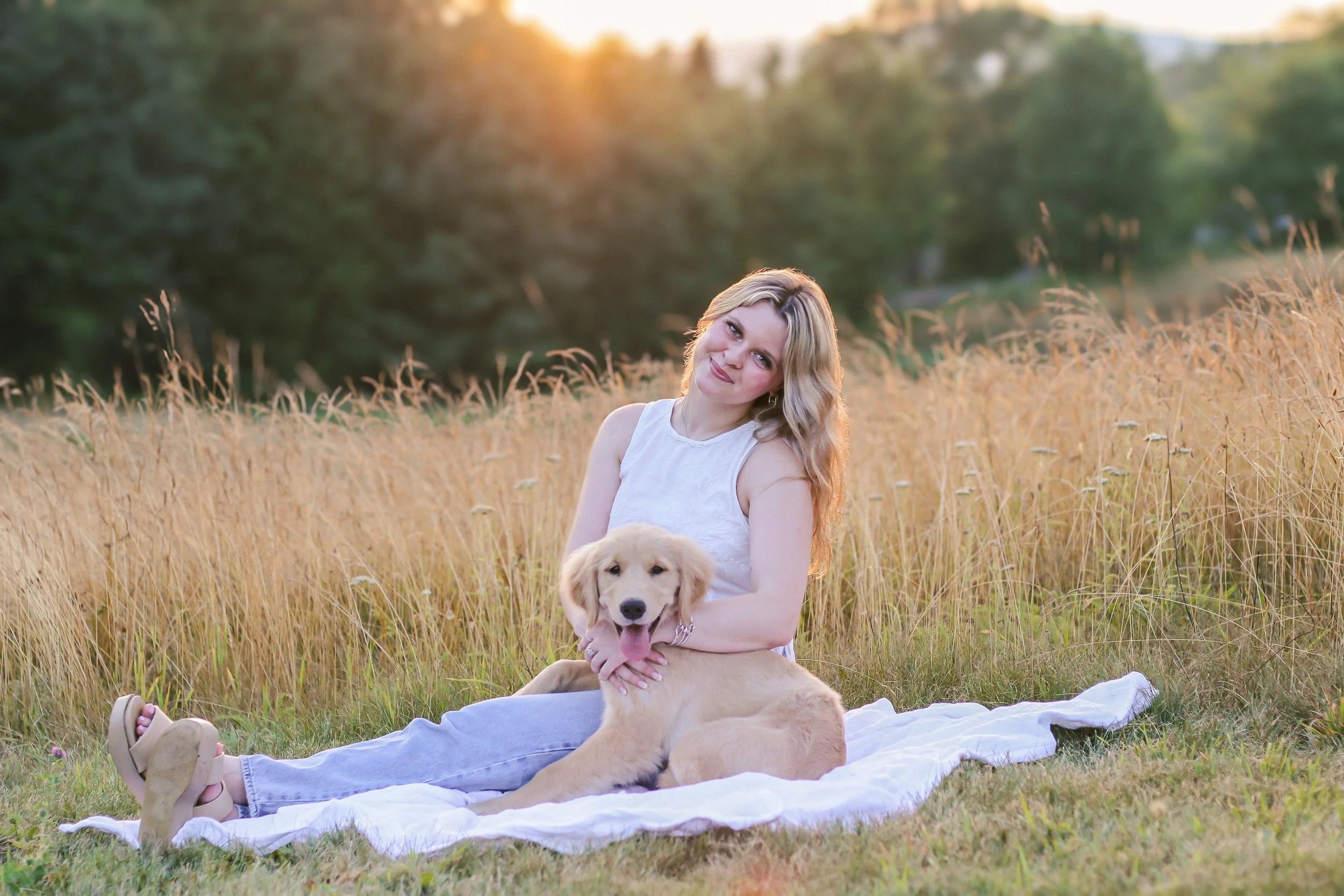 A woman with blonde hair sitting on a white blanket in a field during sunset, holding a golden retriever puppy.