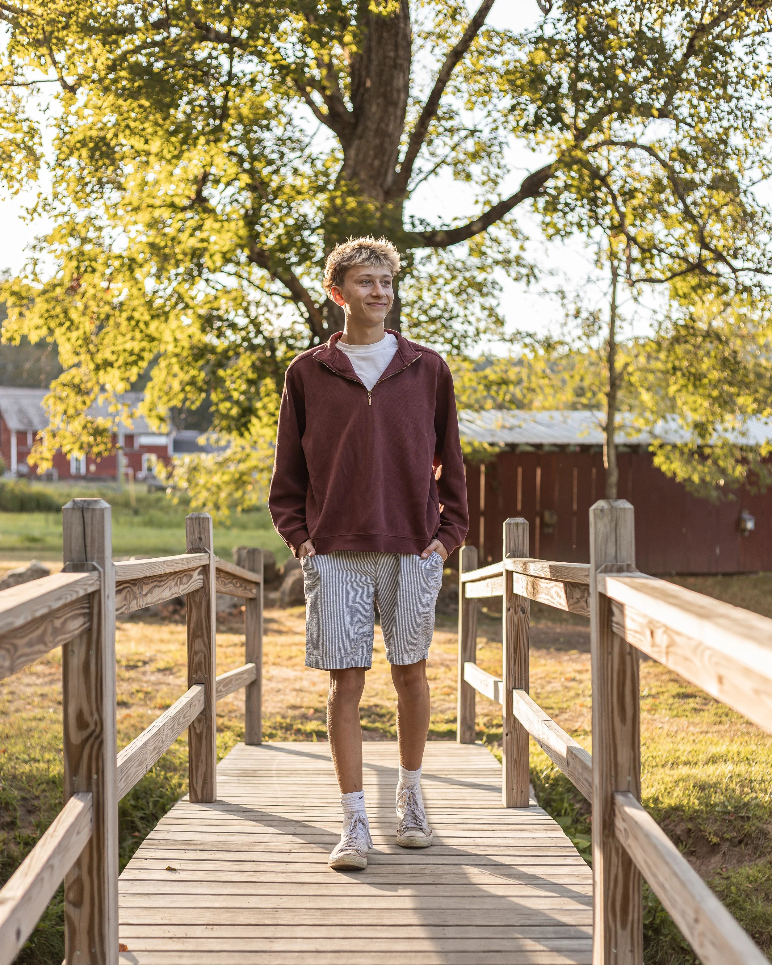 A young man walking on a wooden bridge outdoors during daylight with trees and a barn in the background.