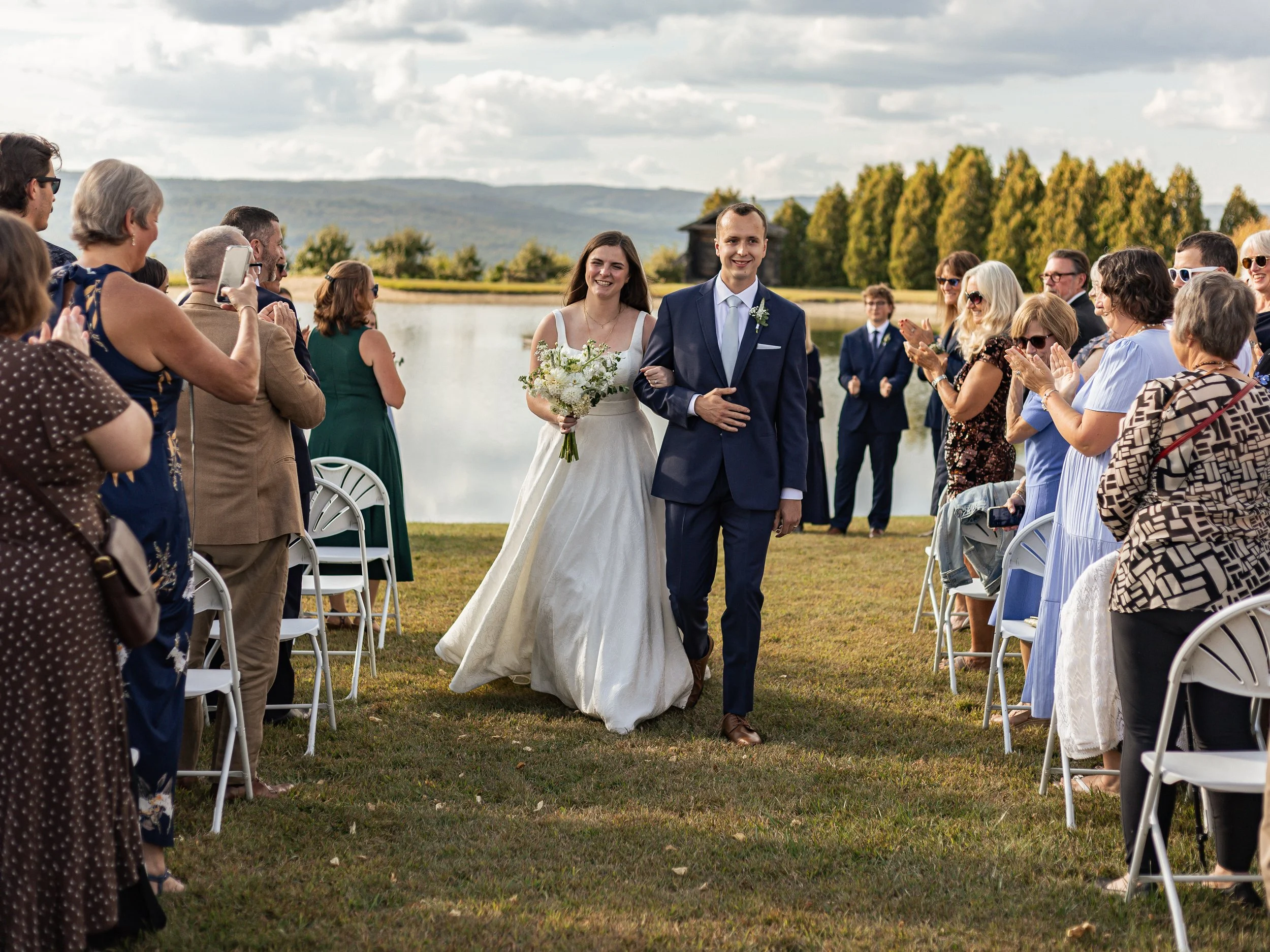 A newlywed couple walking down the aisle at an outdoor wedding, with wedding guests clapping and taking photos by a lake and trees under a partly cloudy sky.
