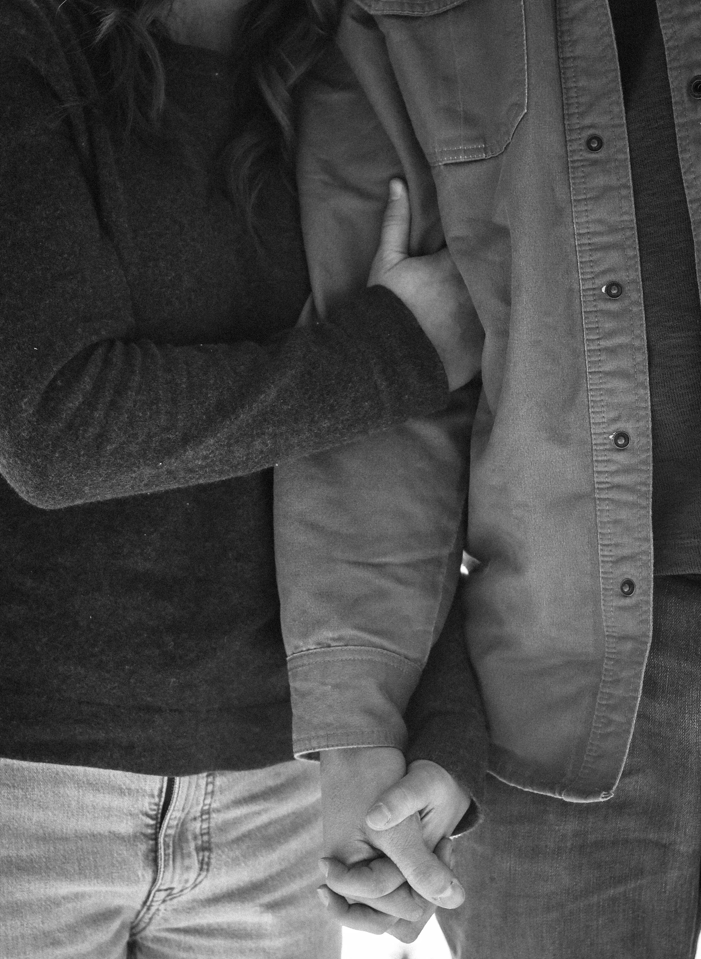 A black and white photo of two people holding hands. One person's hand rests on the other's chest, with their fingers interlinked, and the other person's hand is clasped with the first, with both hands partially visible.