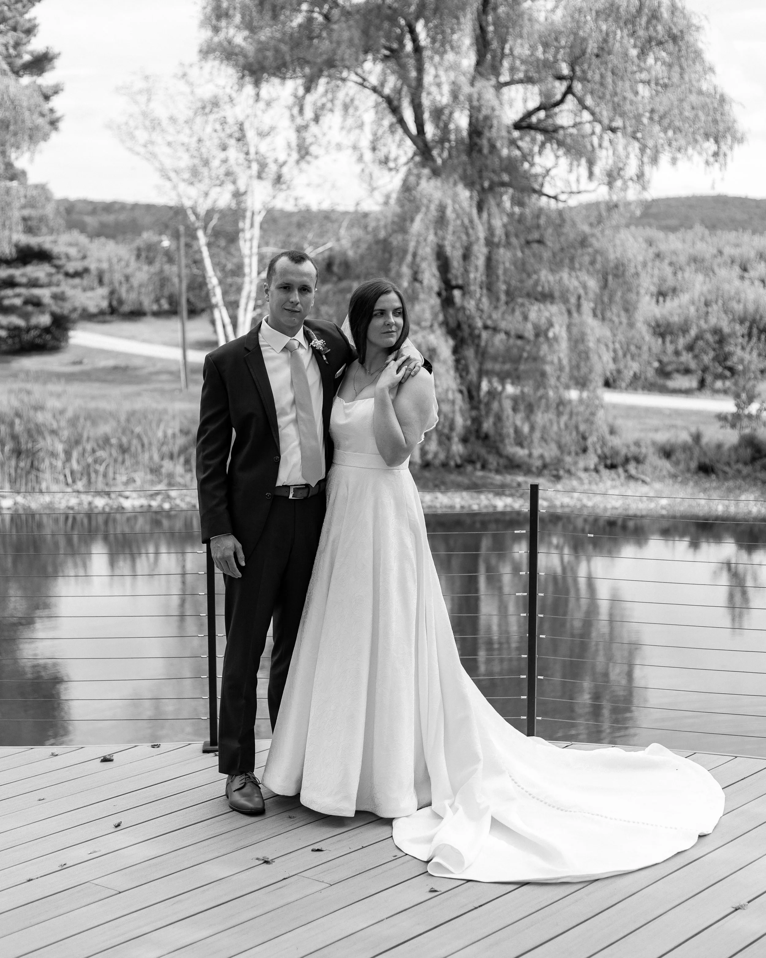 A black and white photo of a bride and groom standing on a wooden deck near water, with trees and hills in the background. The groom wears a suit, and the bride wears a white wedding dress. They stand close together, with the bride resting her hand o