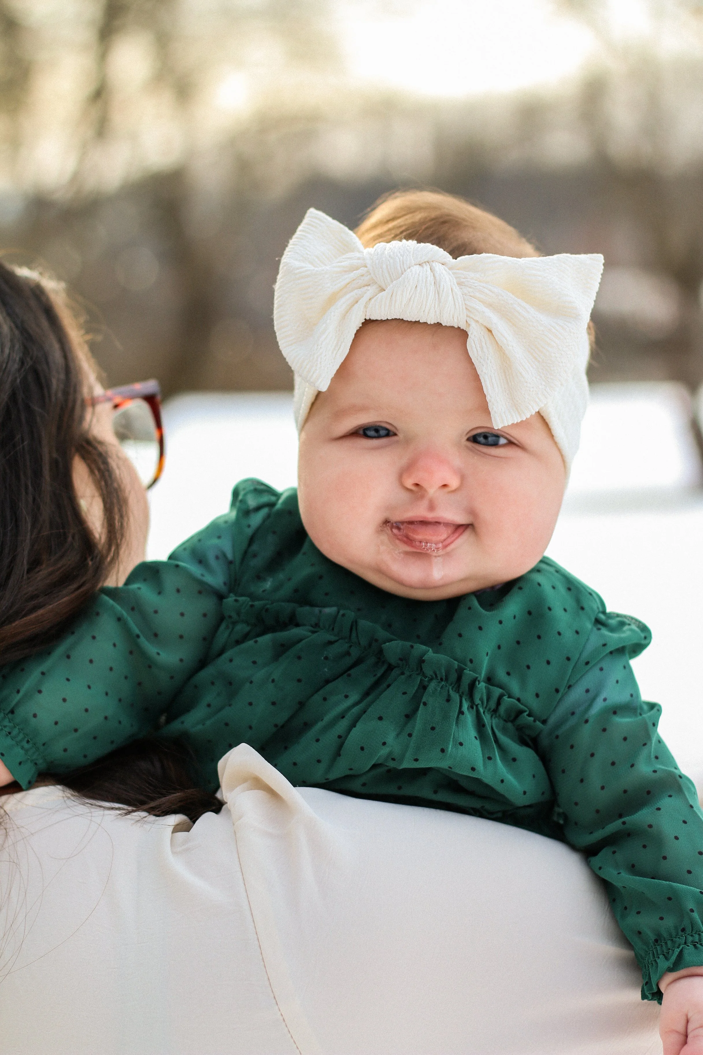 A baby girl wearing a green polka dot dress and a cream-colored headband with a bow, sitting on an adult's shoulder outdoors during winter.