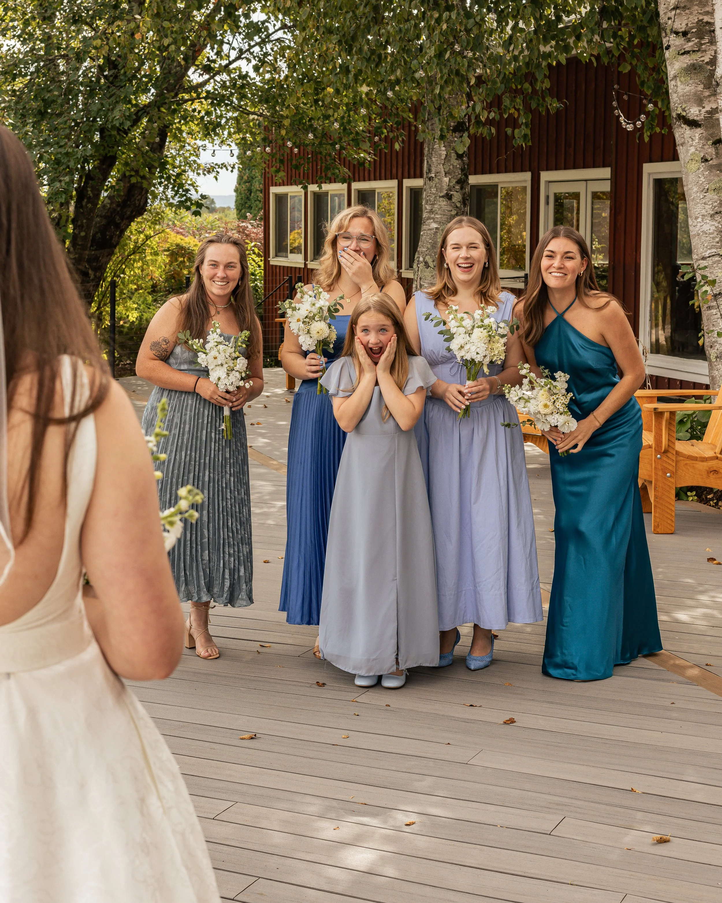 A group of five women and one girl standing outdoors on a wooden deck, smiling and holding bouquets at a wedding or celebration, with trees and a red building in the background.