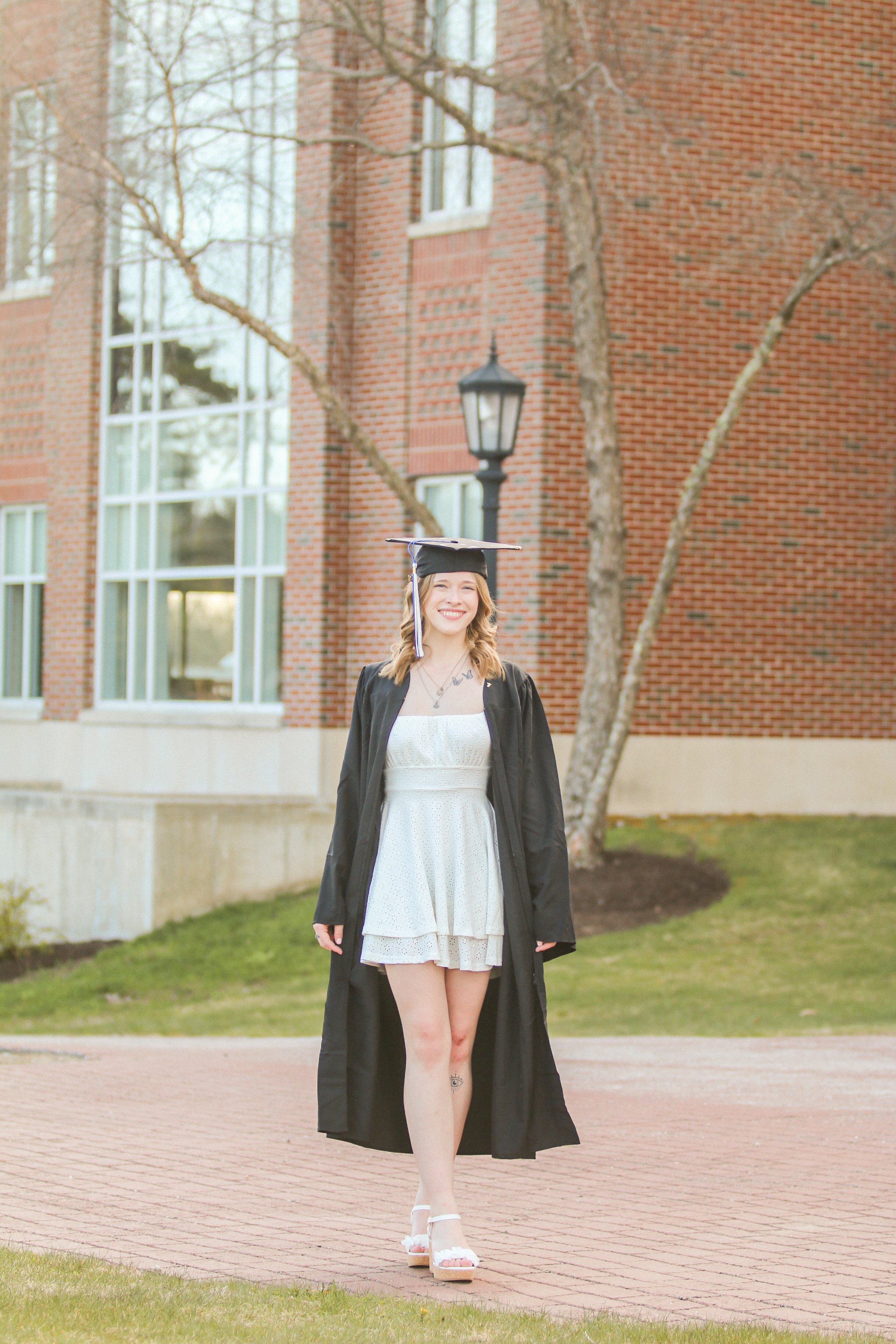 A young woman in a graduation cap and gown standing outdoors in front of a brick building, smiling.