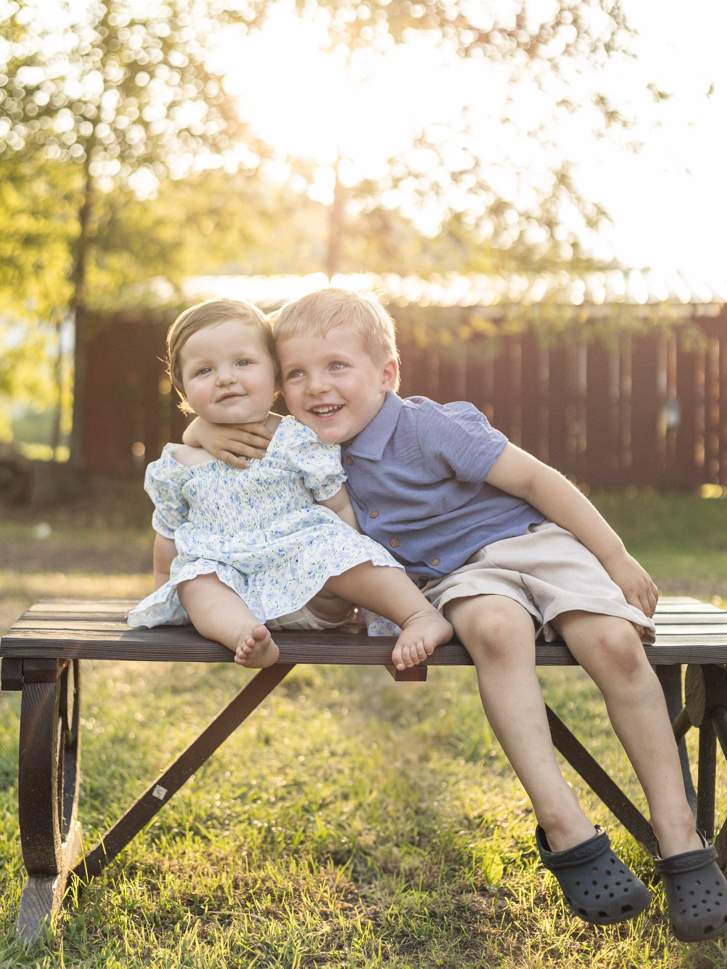 A young boy and a young girl sitting on a park bench outside during sunset, hugging and smiling.