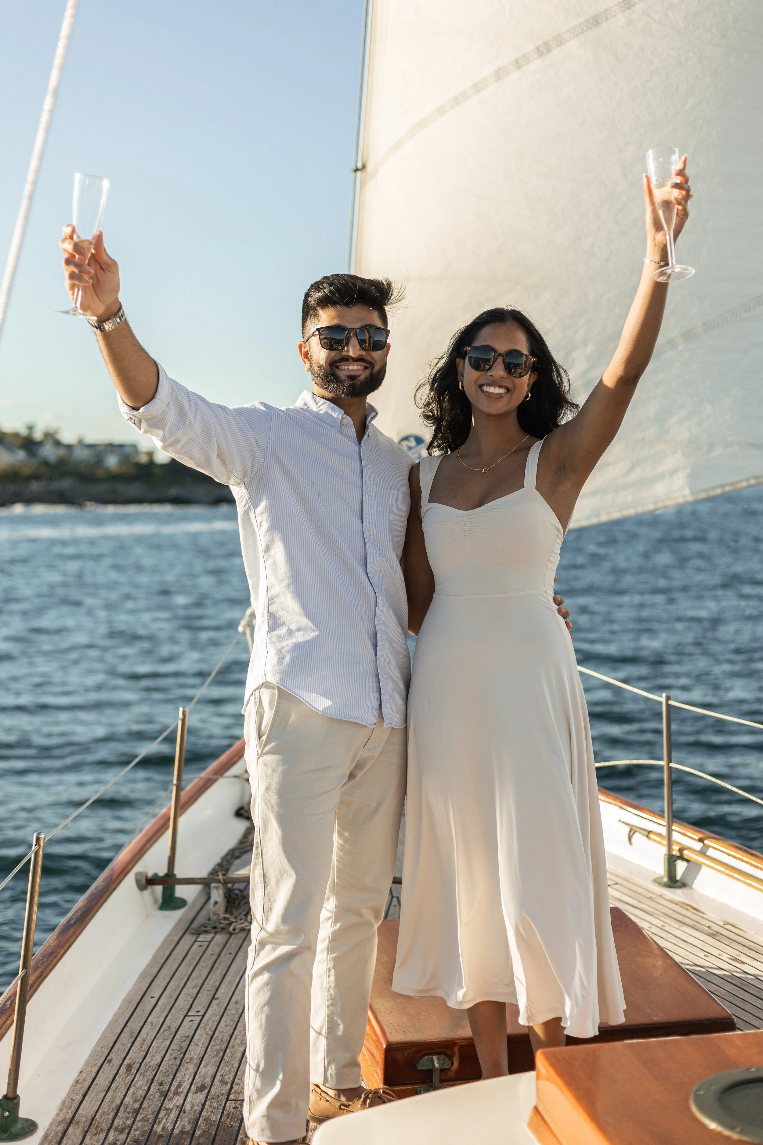 A smiling couple standing on a sailboat, raising champagne glasses in celebration. The man is wearing a light-colored shirt and pants, and the woman is in a white dress. Both are wearing sunglasses, with water and a sail in the background.