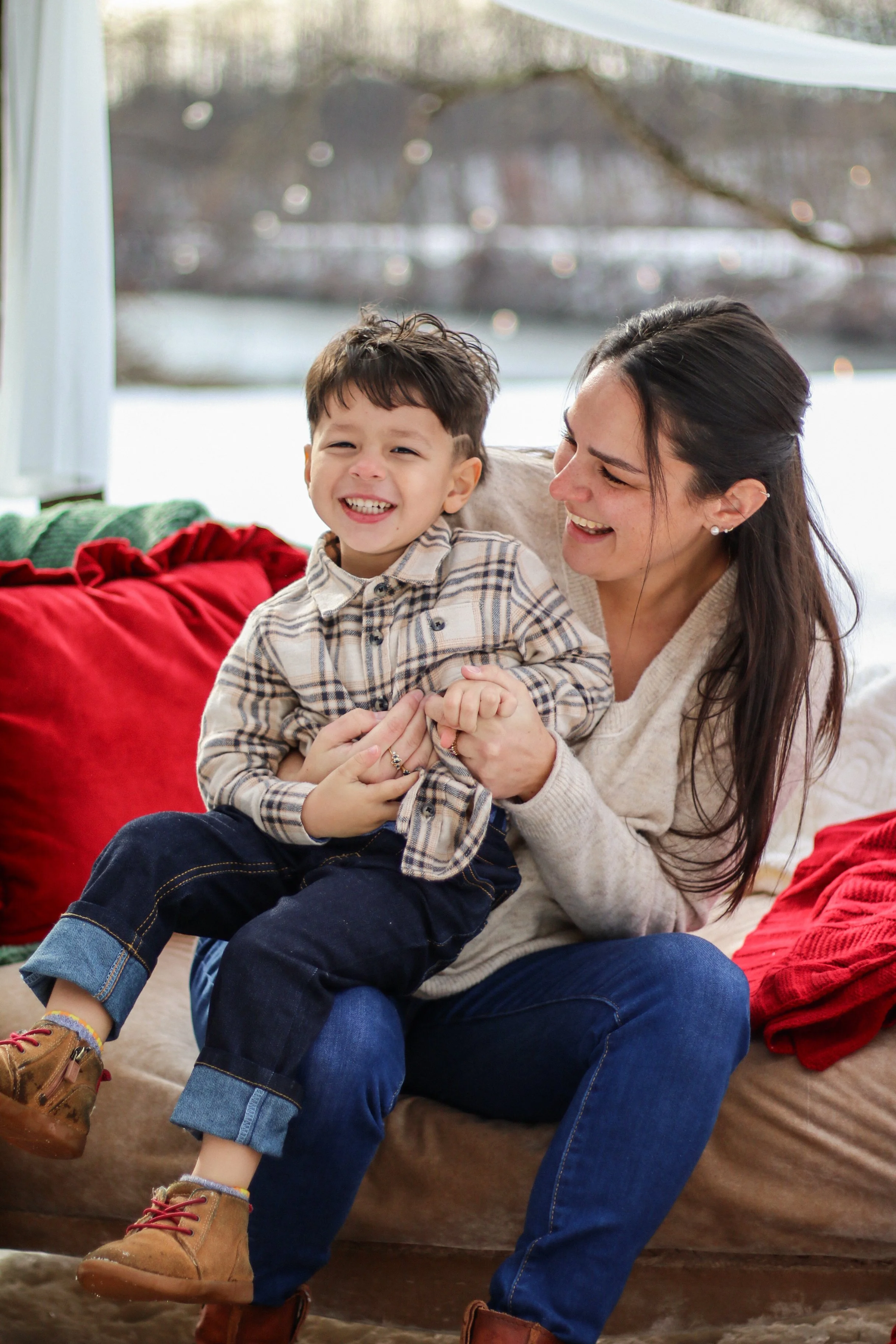 A woman and young boy laughing and playing together on a beige sofa with red pillows, in front of a snowy windowed background.