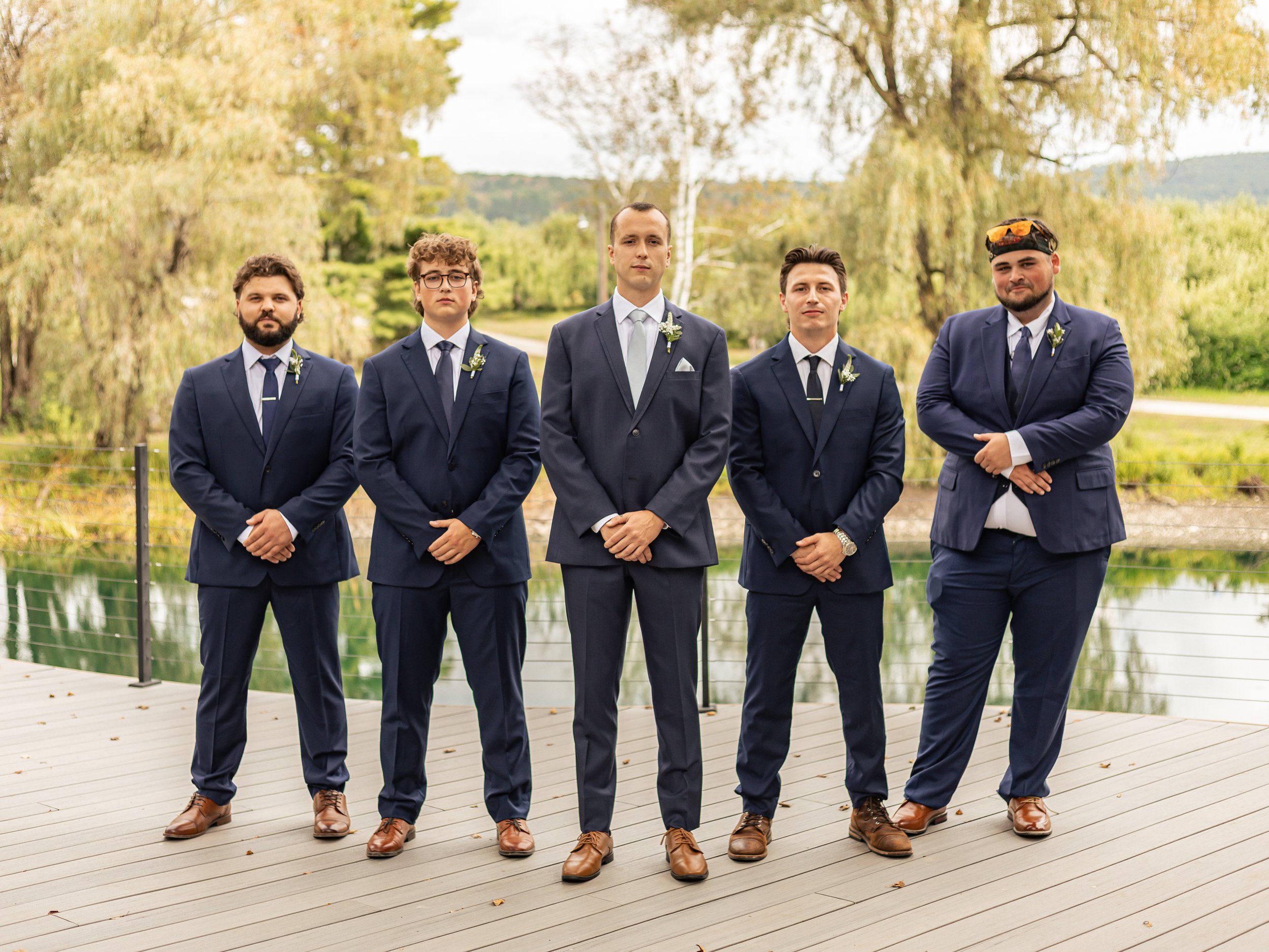 Five men dressed in navy suits standing outdoors on a wooden deck near a body of water, with trees and greenery in the background.
