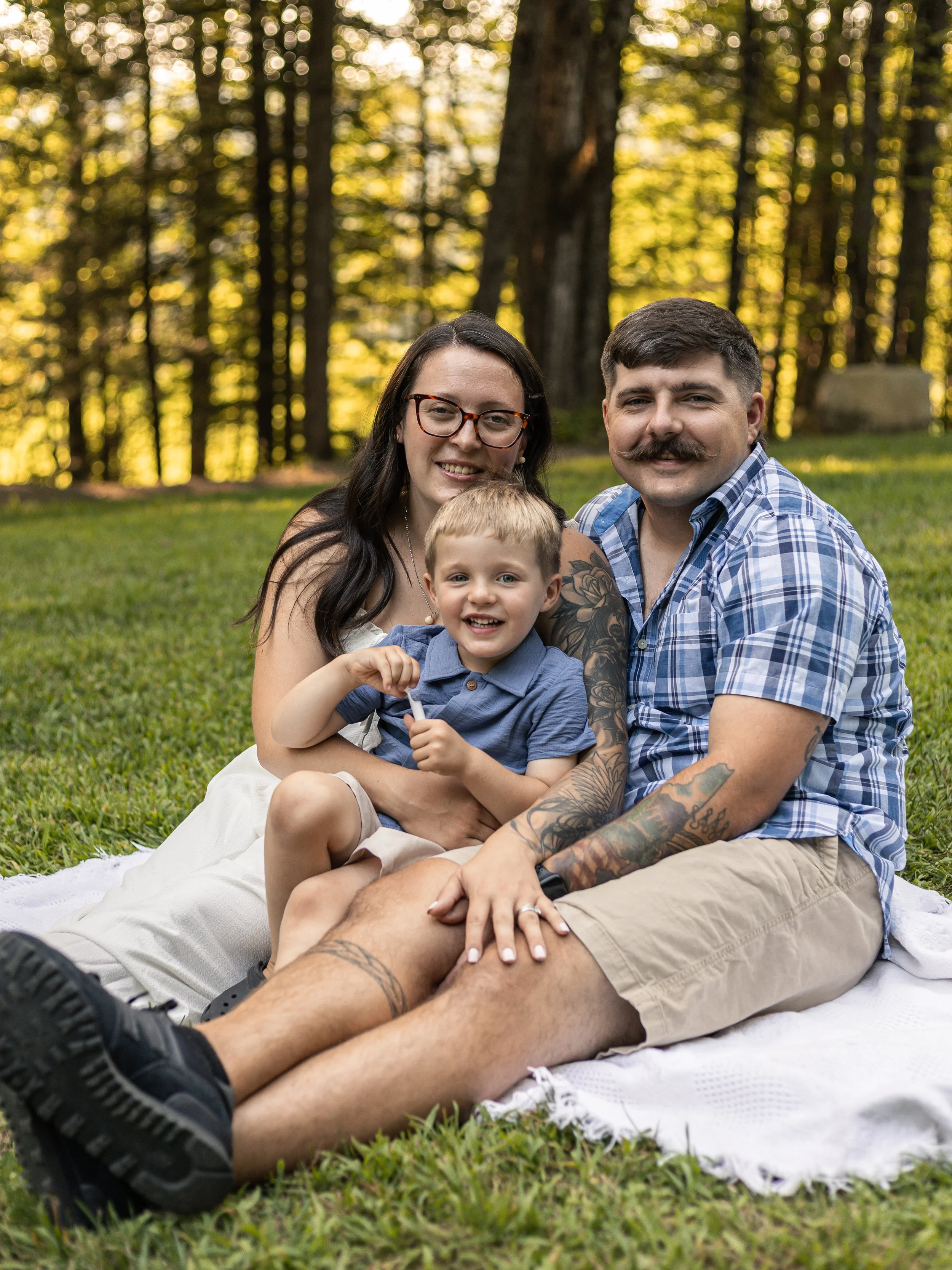 A family of three sitting on a blanket outdoors in a park with trees in the background during late afternoon or early evening.