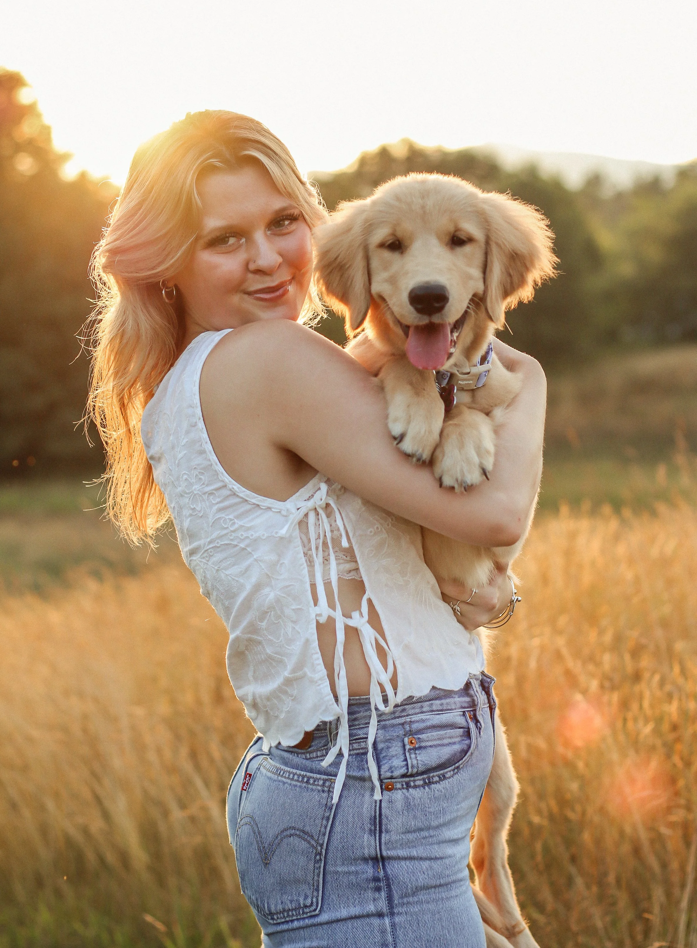 A woman in a white sleeveless top holding a golden retriever puppy in a field during sunset.