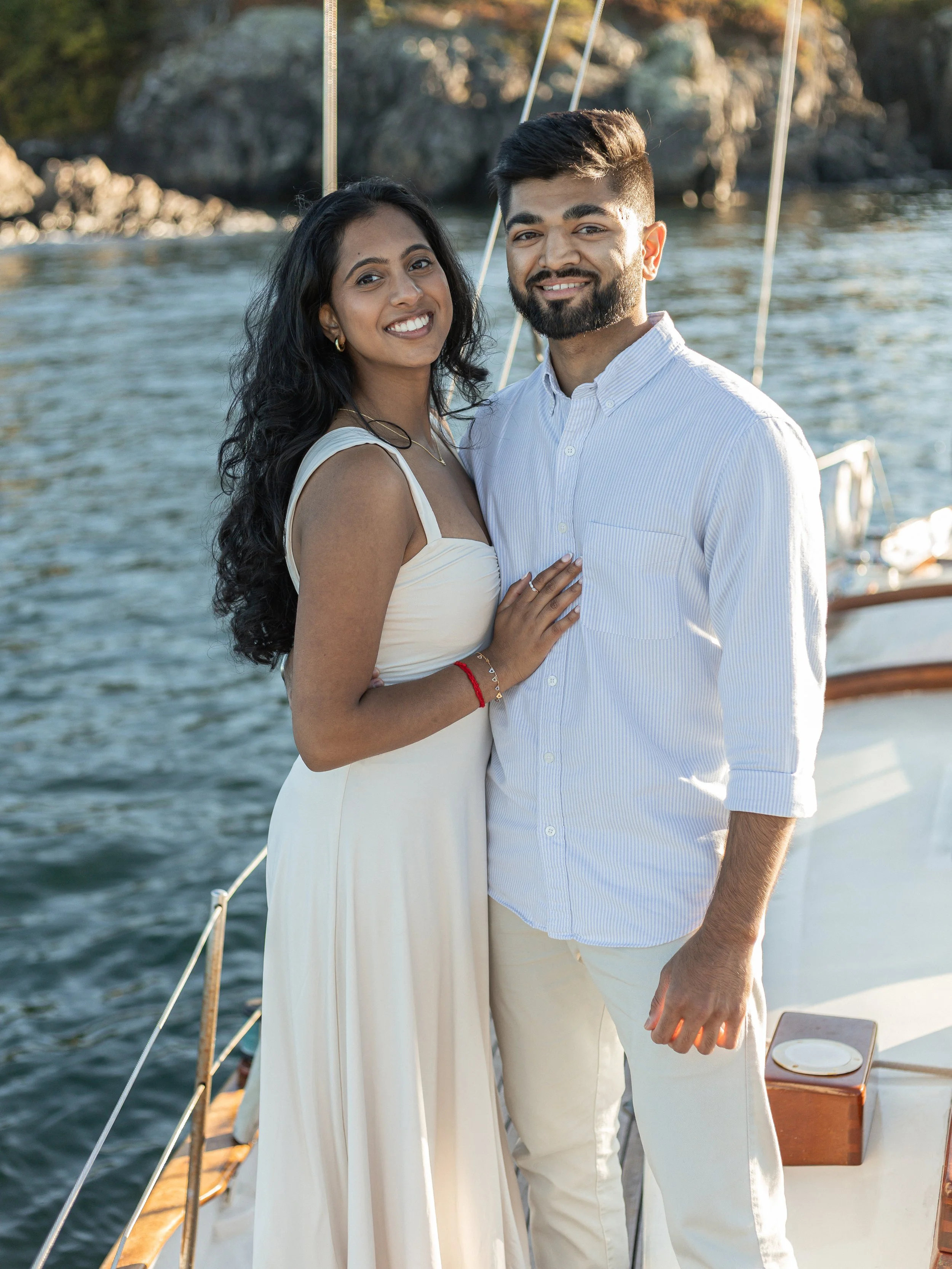 A smiling woman and man standing close together on a boat, with water and rocks in the background.