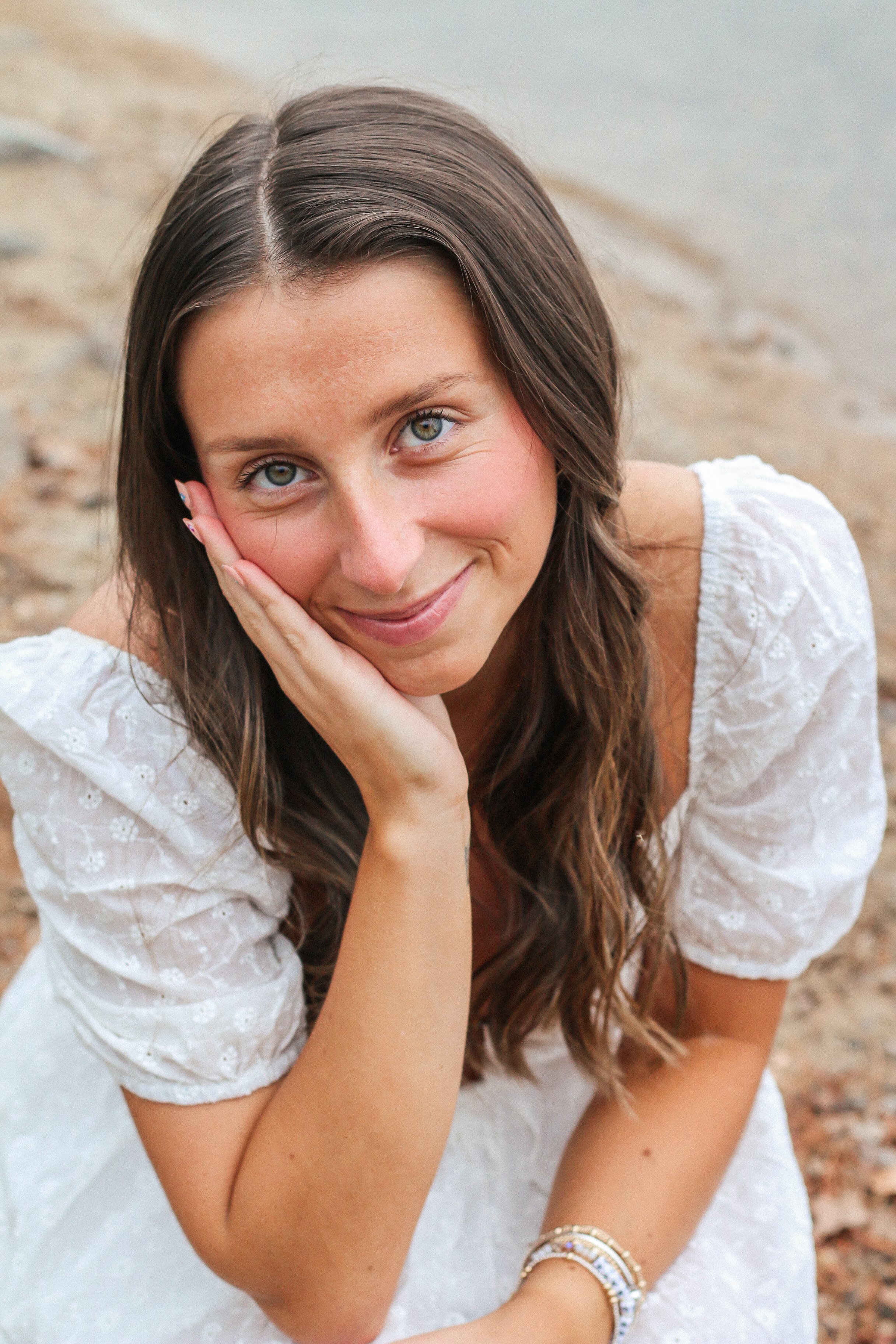 A young woman with long brown hair and blue eyes smiling softly, resting her head on her left hand, sitting outdoors on sandy ground.
