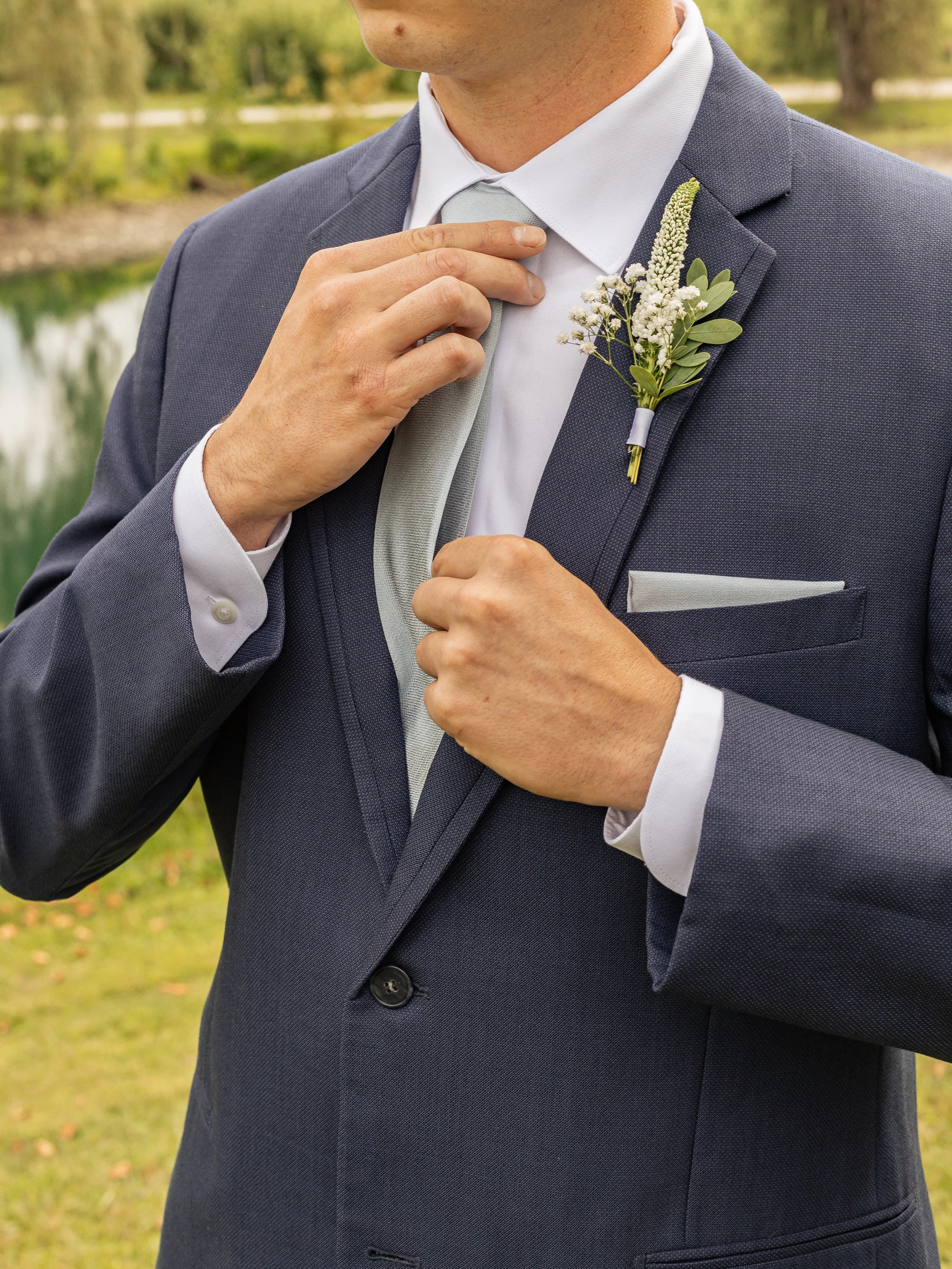 A man in a dark suit, white dress shirt, and light gray tie adjusting his collar, with a boutonniere of white flowers and green leaves on his lapel, standing outdoors near a body of water and trees.