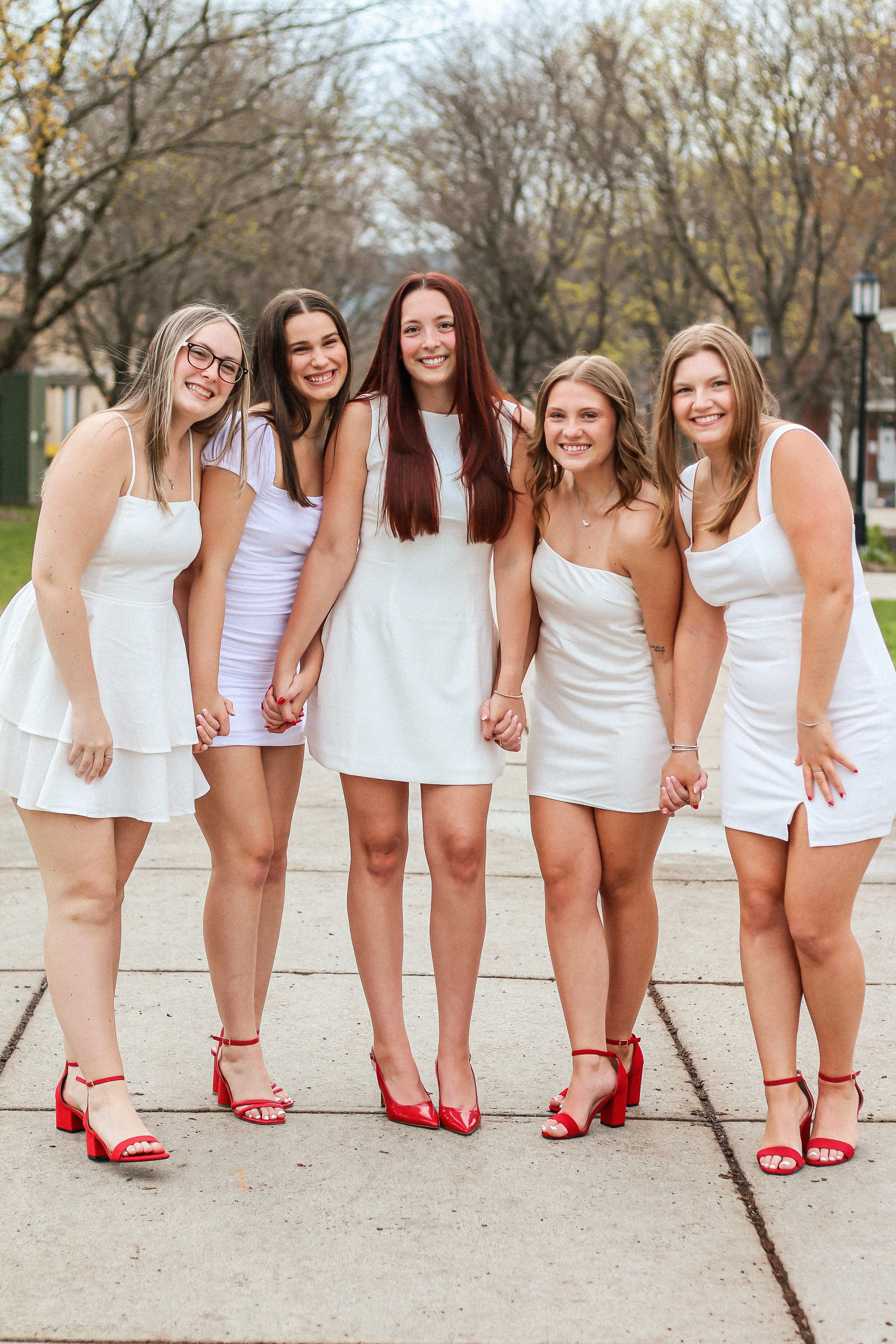Group of five young women dressed in white dresses and red heels, standing outdoors on a sidewalk holding hands and smiling.