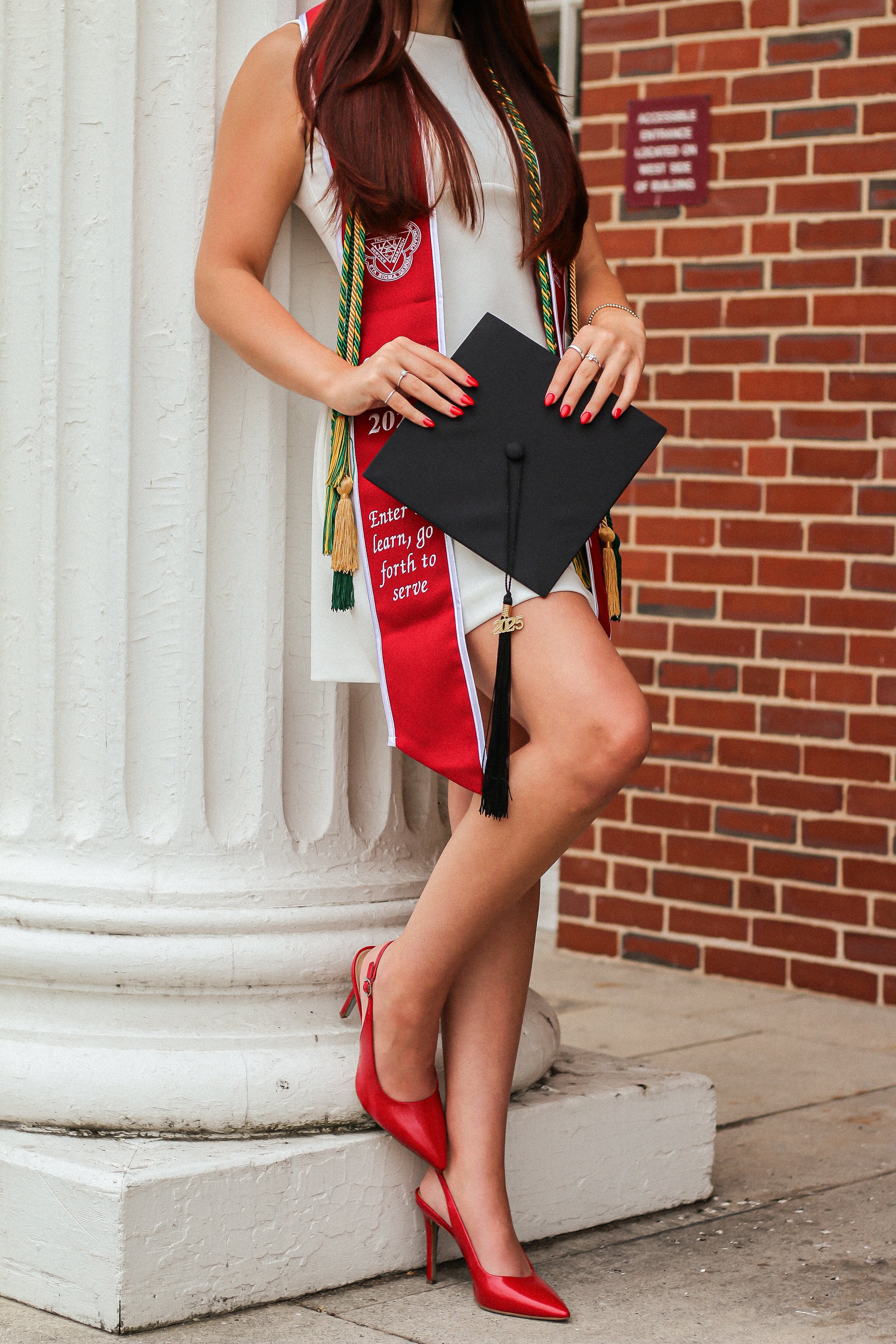 A woman in a white dress with red high heels holding a graduation cap, wearing a red graduation sash with embroidered text, standing in front of a brick wall and a white column.