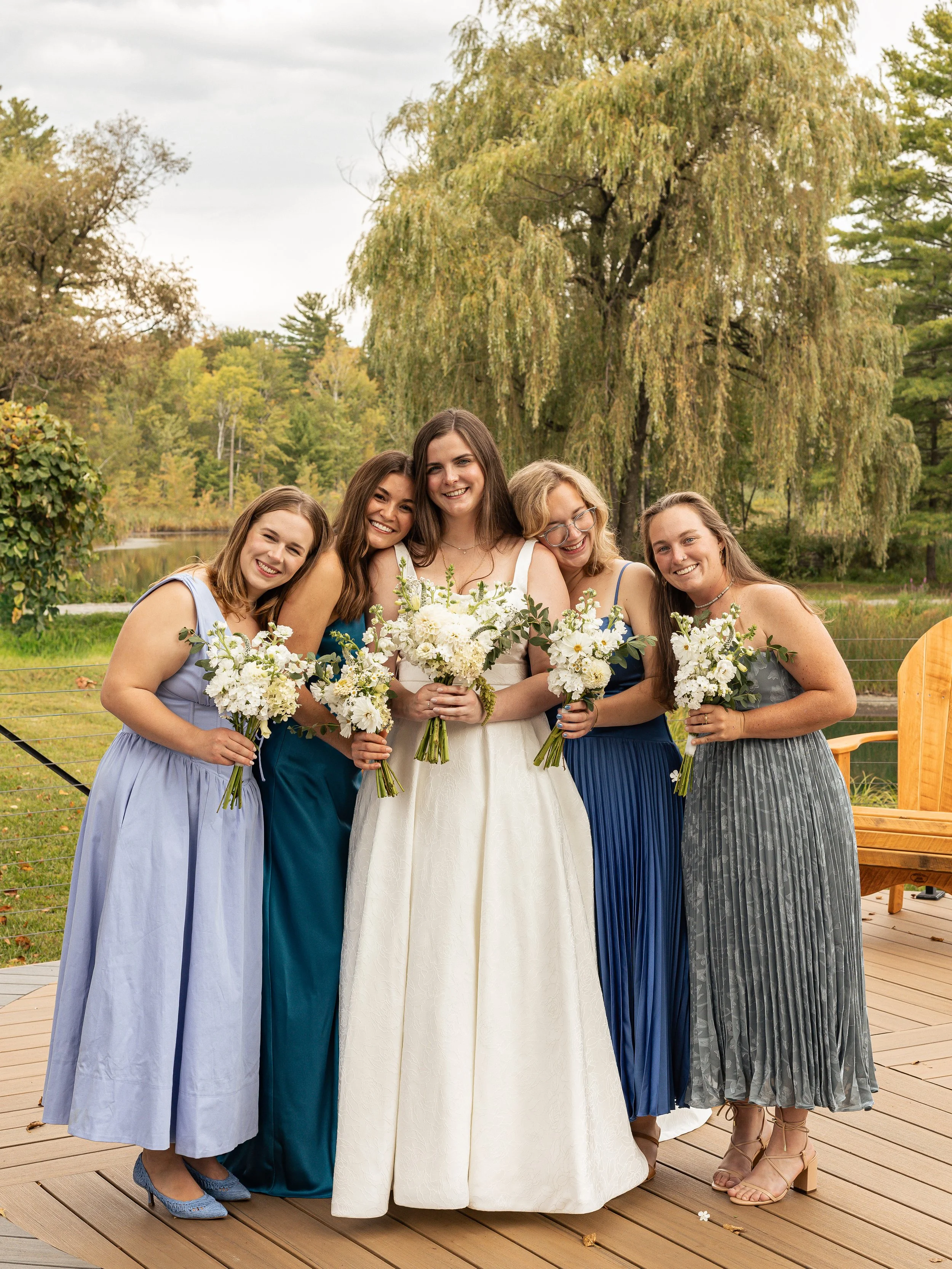 A bride in a white dress with four bridesmaids in colorful dresses, holding white flower bouquets, outside on a wooden deck with trees and a pond in the background. 