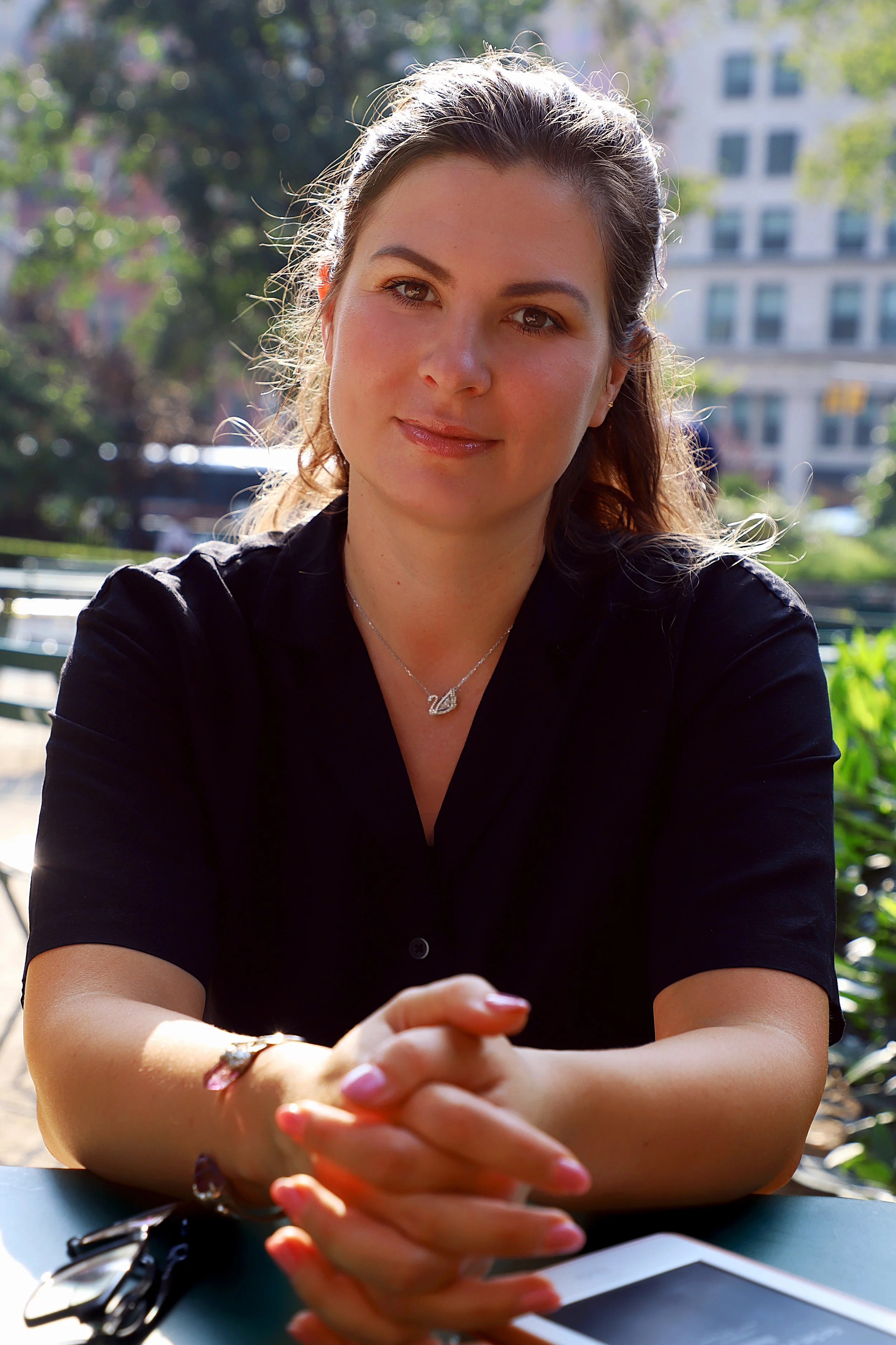 Young woman sitting outdoors at a table with sunlight on her face, wearing a black shirt and silver necklace, with trees and a building in the background.