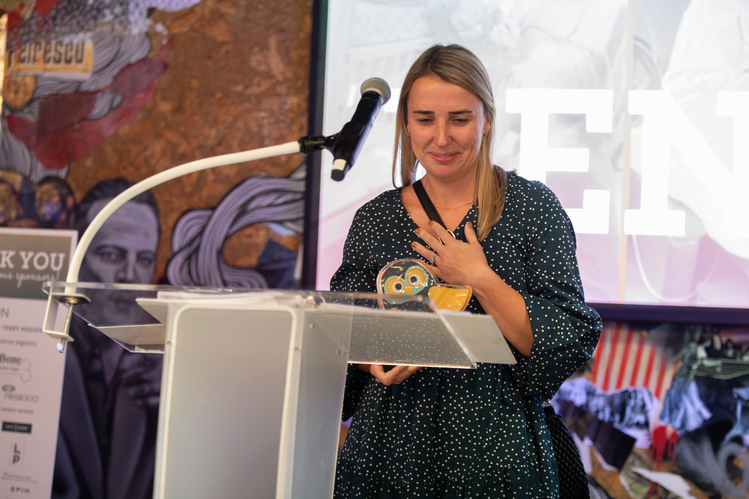 Woman holding an award at a podium with microphone, smiling in front of a colorful mural background.