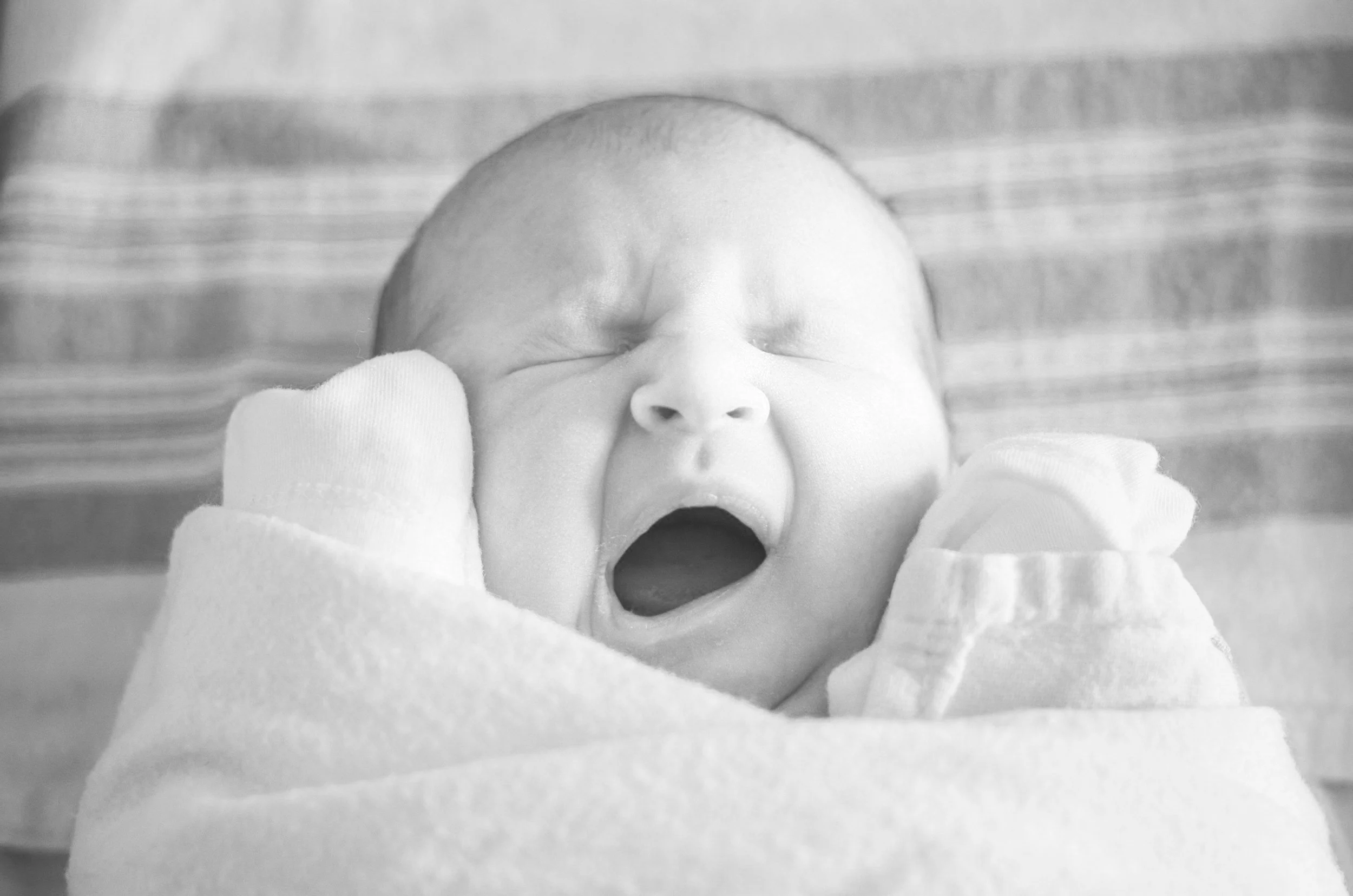 Black and white photo of a newborn baby yawning, wrapped in a blanket.
