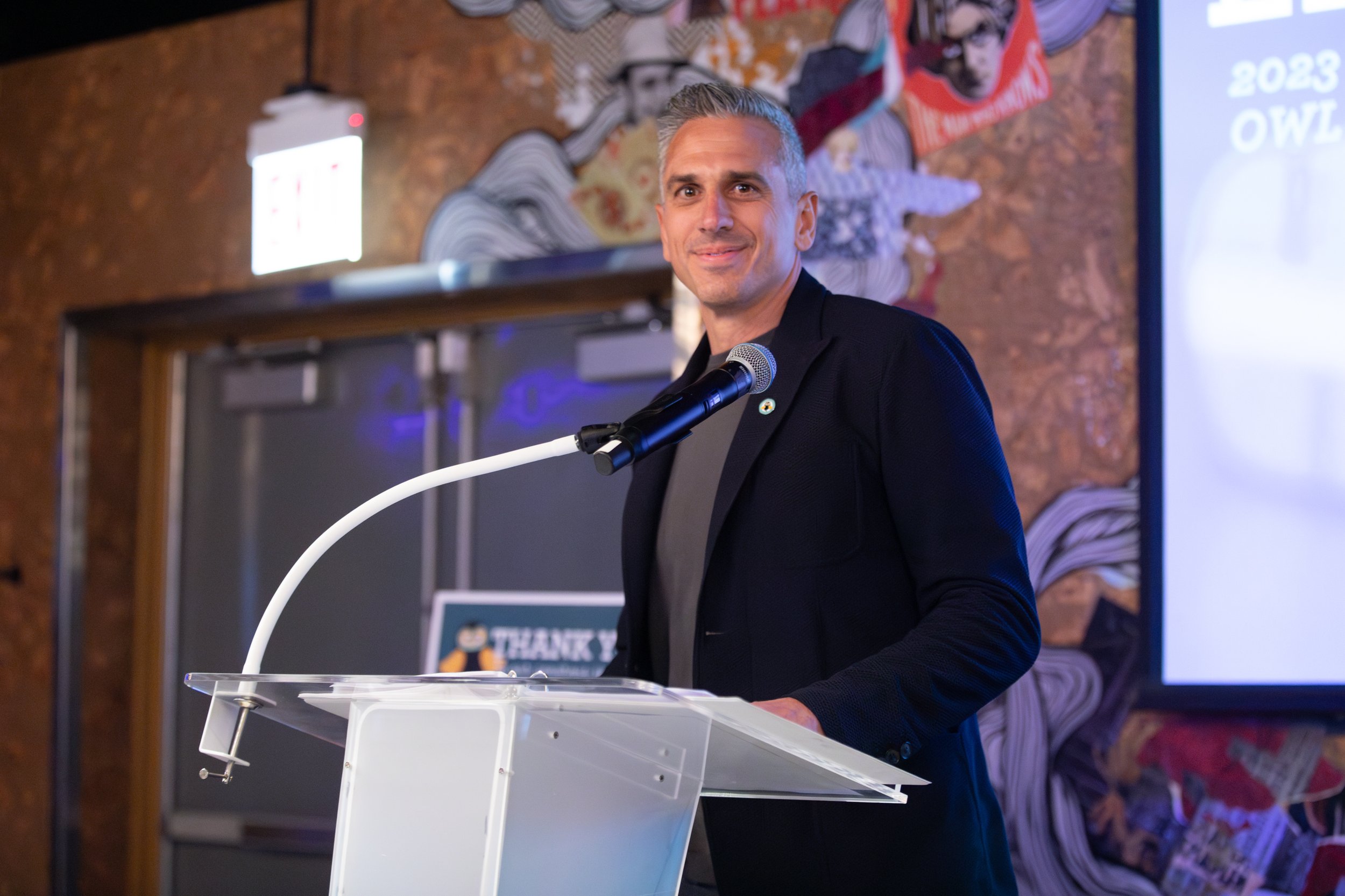 Person speaking at a podium with a microphone, wearing a suit, in front of a colorful background and exit sign.