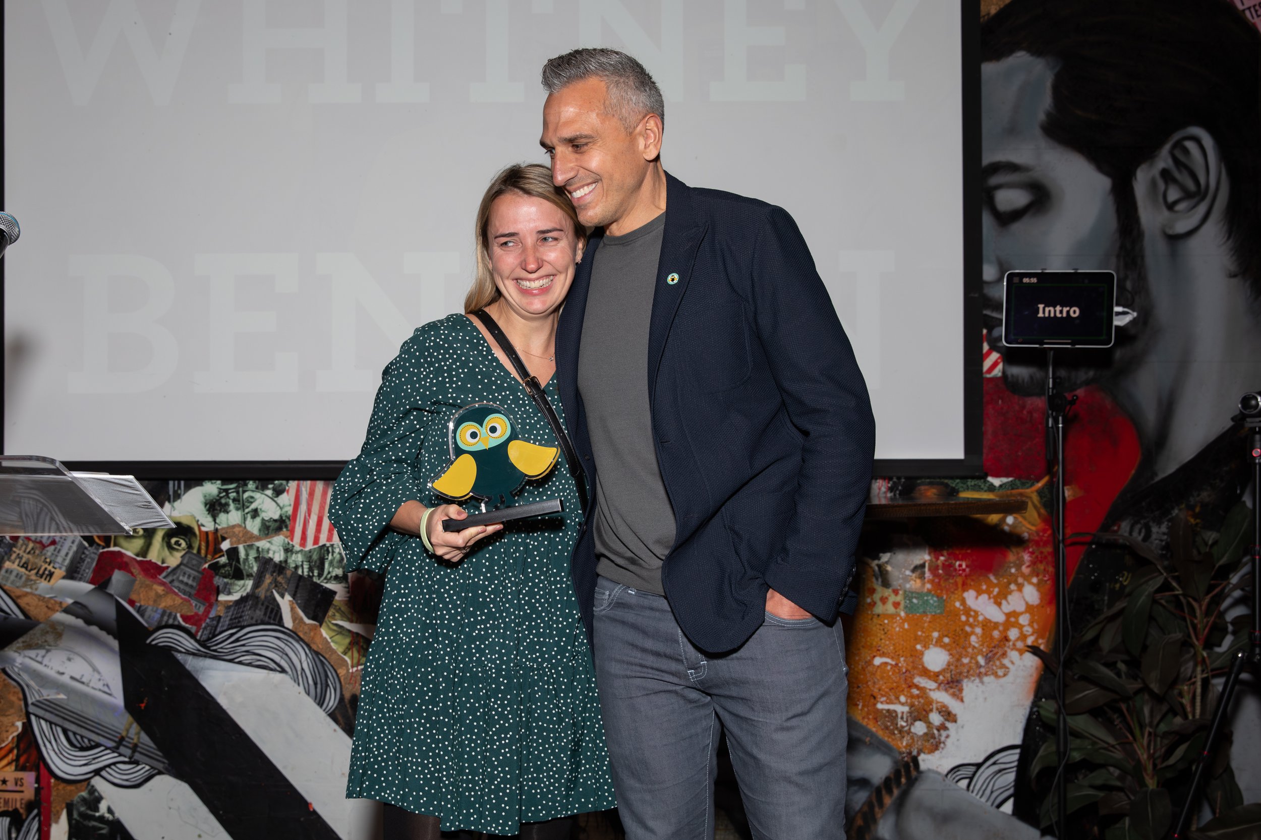 Two people smiling on stage, one holding an award shaped like an owl. Background features a projector screen and colorful artwork.