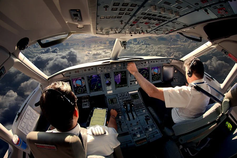 Cockpit of an airplane in flight, with two pilots looking at instruments and the sky visible through the windshield.
