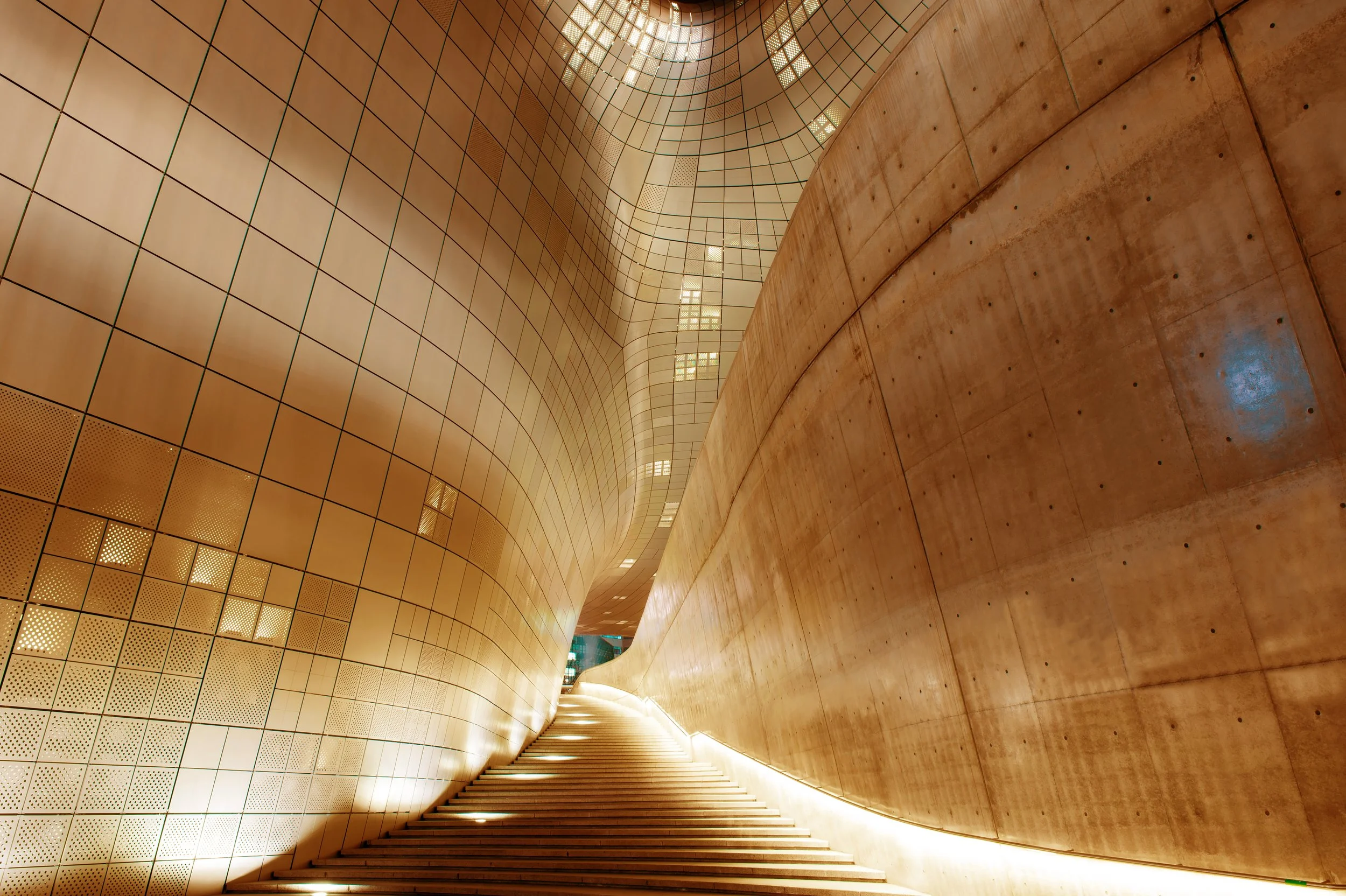 The image shows an interior view of a modern architectural structure with curved walls, a staircase, and high ceilings with glass windows, featuring warm lighting and a combination of wood and tiled surfaces.