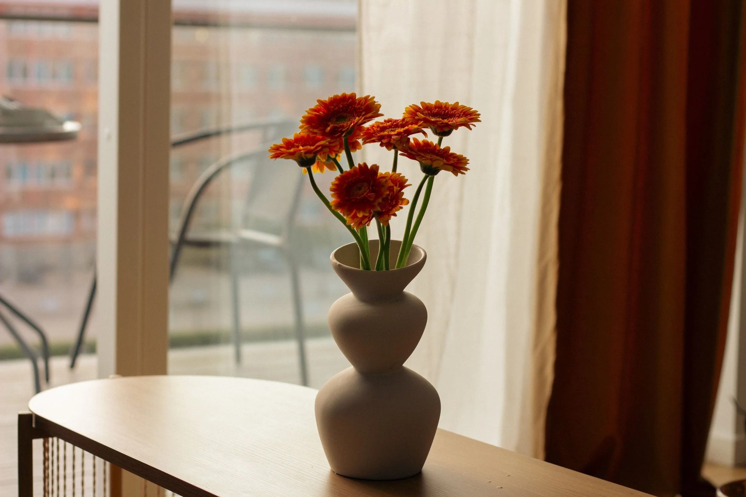 A bouquet of orange-red flowers in a white ceramic vase on a wooden table, near a window with curtains, overlooking a balcony with a chair outside.