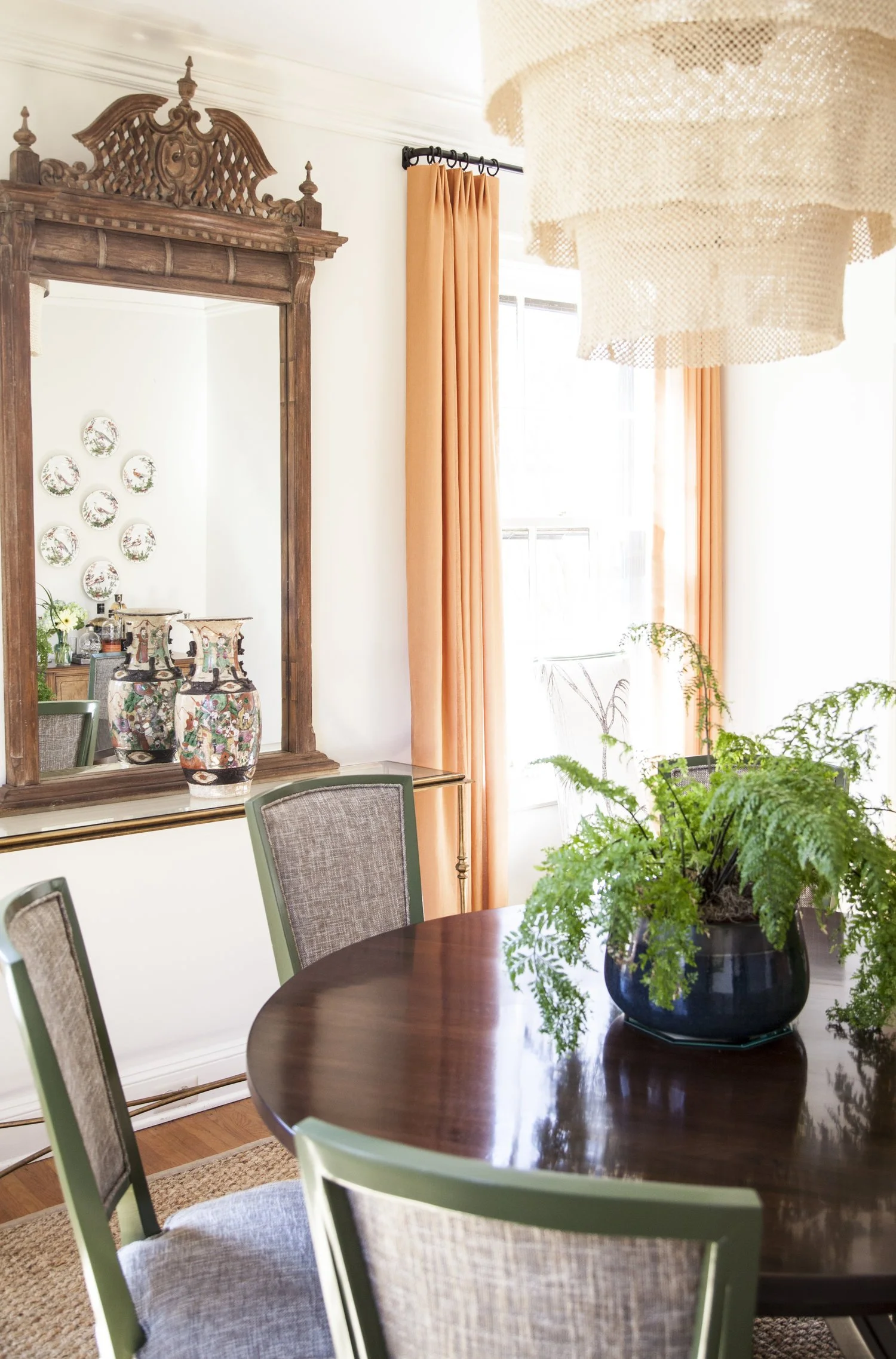 K. Interiors Design Work - A dining room with a wooden table, upholstered chairs, a large plant in a blue pot, a antique mirror, colorful vases, and orange curtains by a window.