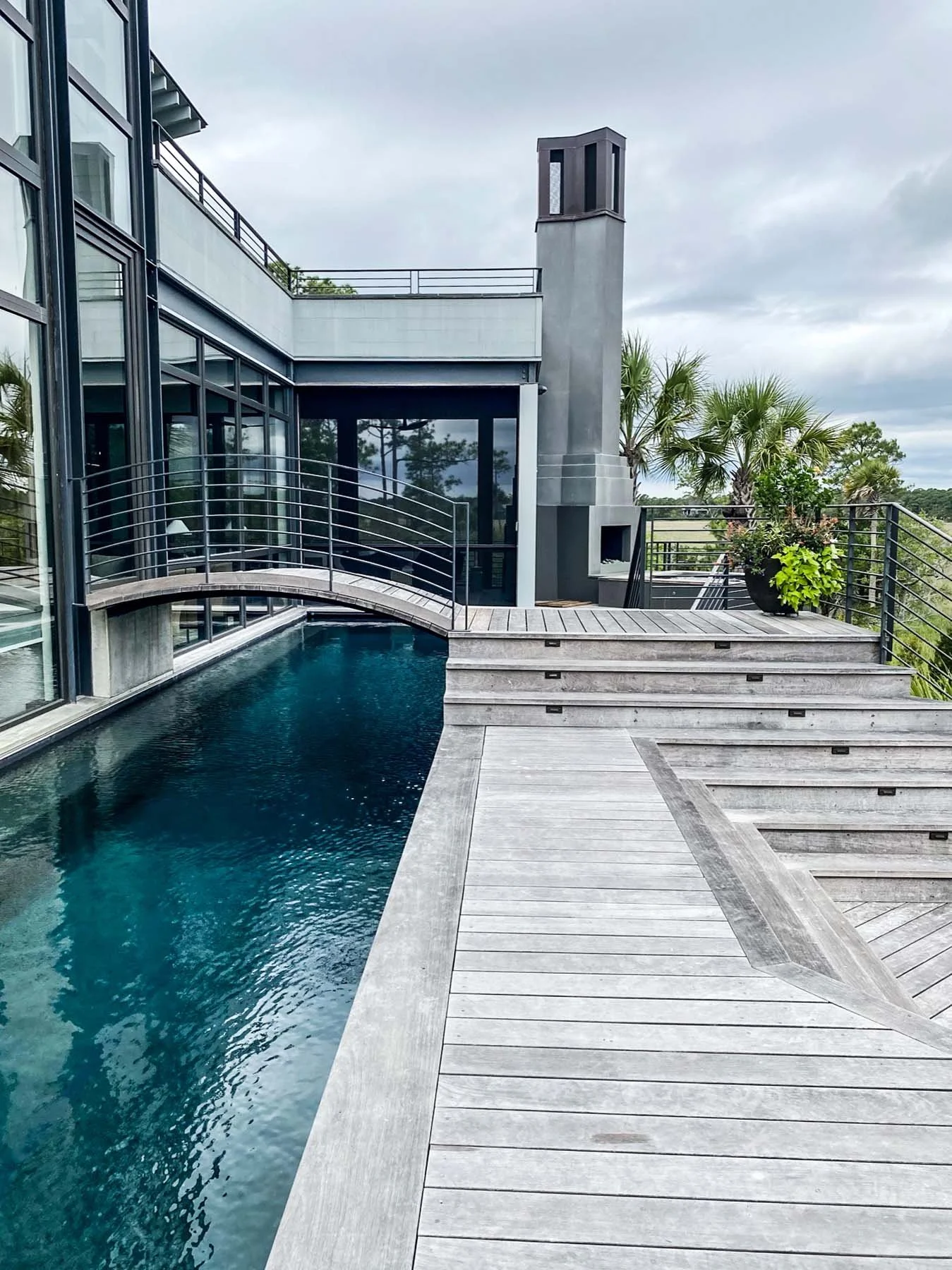 K. Interiors Design Work - Modern house with outdoor swimming pool, wooden deck, and railing, overlooking palm trees on cloudy day.