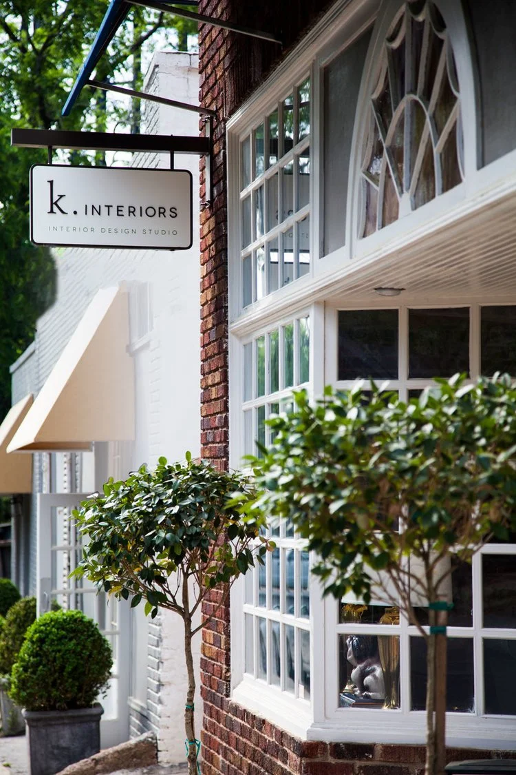 Exterior of a building with a sign reading 'k. INTERIORS Interior Design Studio', large windows, potted trees, and a sidewalk.
