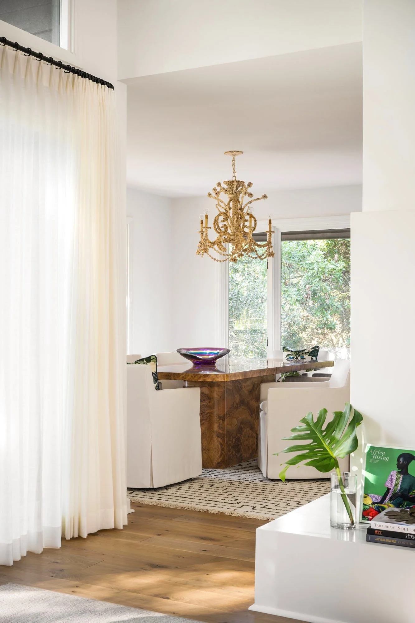 K. Interiors Design Work - Dining area with a wooden table, white chairs, a gold chandelier, and a large window showing trees outside.