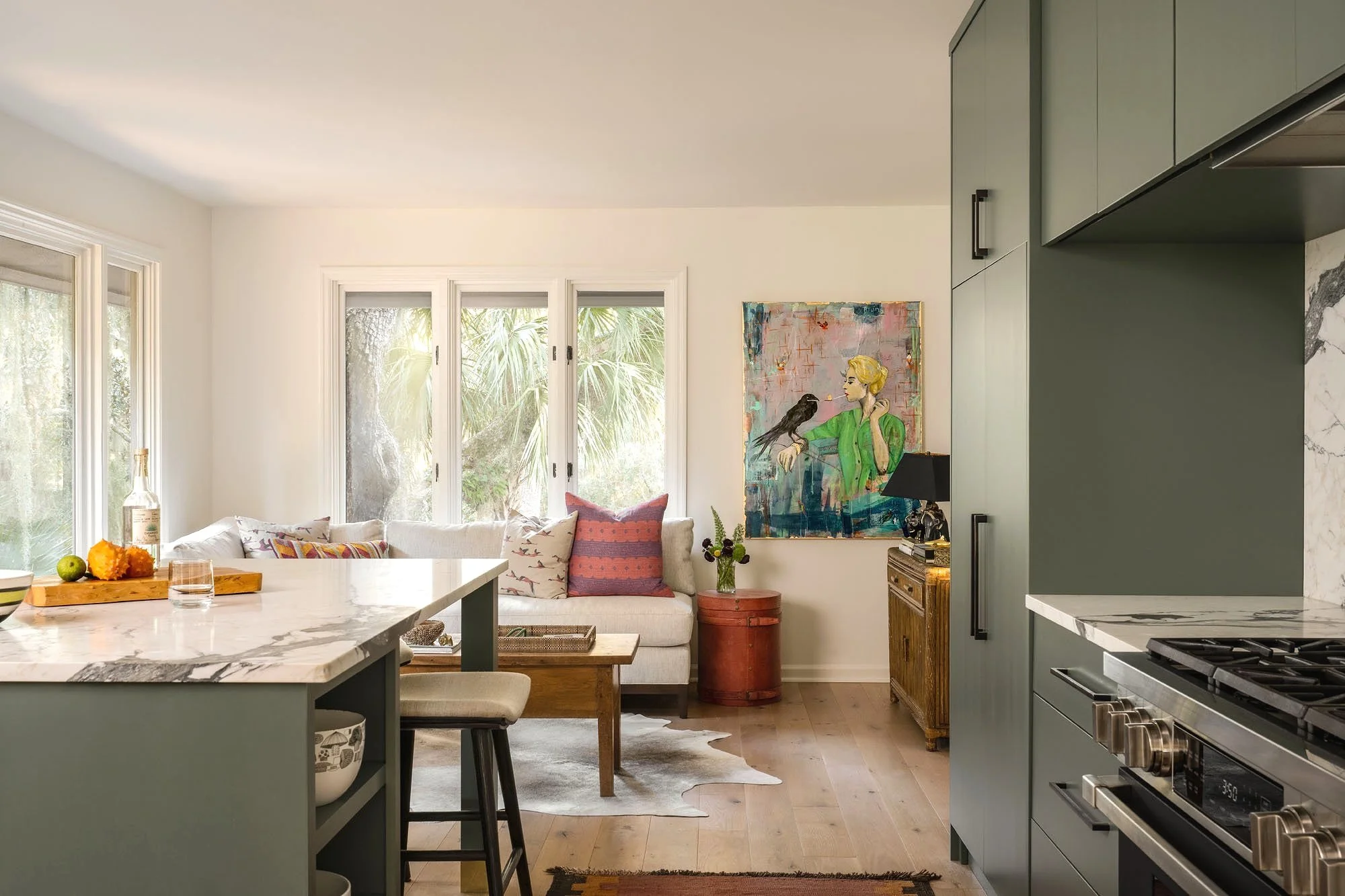 K. Interiors Design Work - A bright living room with large windows, a white sofa with colorful pillows, a wooden coffee table, and wall art of a woman with a bird on her arm. Part of a kitchen with green cabinets and a marble countertop is visible.