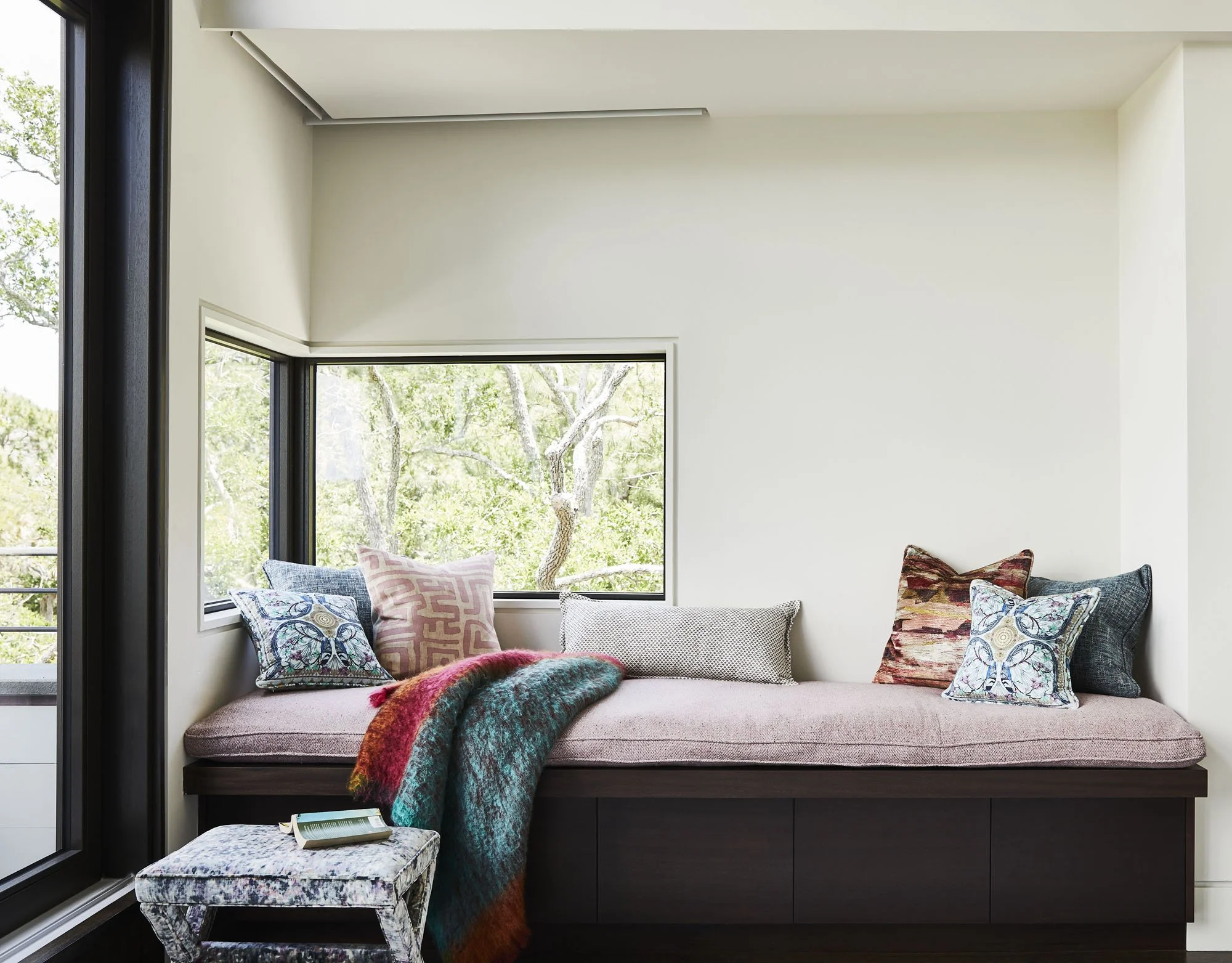 K. Interiors Design Work - Sunlit window nook with a cushioned window seat decorated with colorful pillows and a throw blanket, overlooking leafy trees outside.