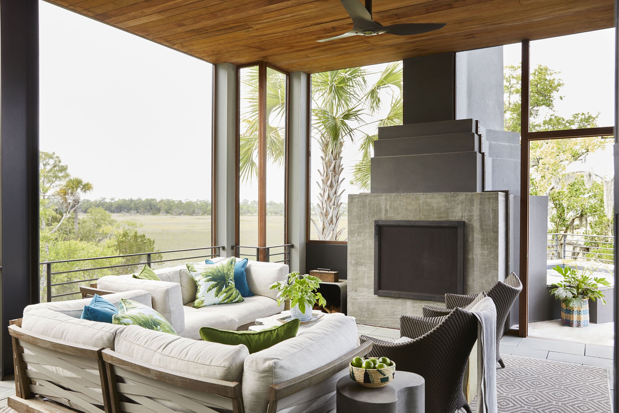 K. Interiors Design Work - Living room with large windows, white sofas with colorful pillows, a concrete fireplace, potted plants, and outdoor scenery visible through the windows.