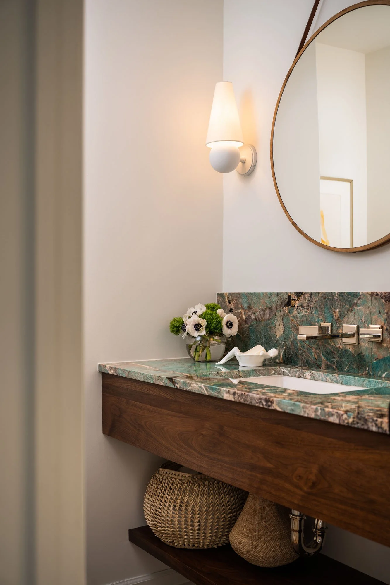 K. Interiors Design Work - Bathroom with a marble countertop, a round mirror, a wall-mounted light, and a vase with white flowers.