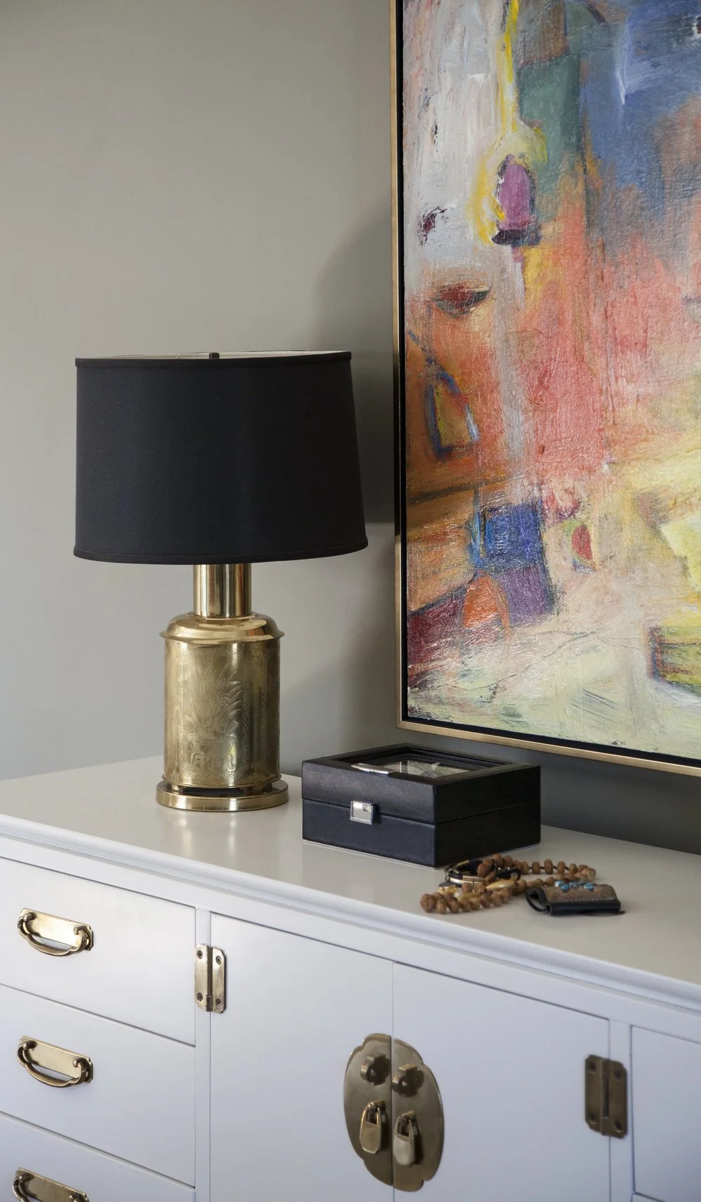 K. Interiors Design Work - A white sideboard with gold handles and a lock, a black and gold lamp, a black jewelry box, and a set of jewelry and beads on top, with an abstract colorful painting on the wall behind.