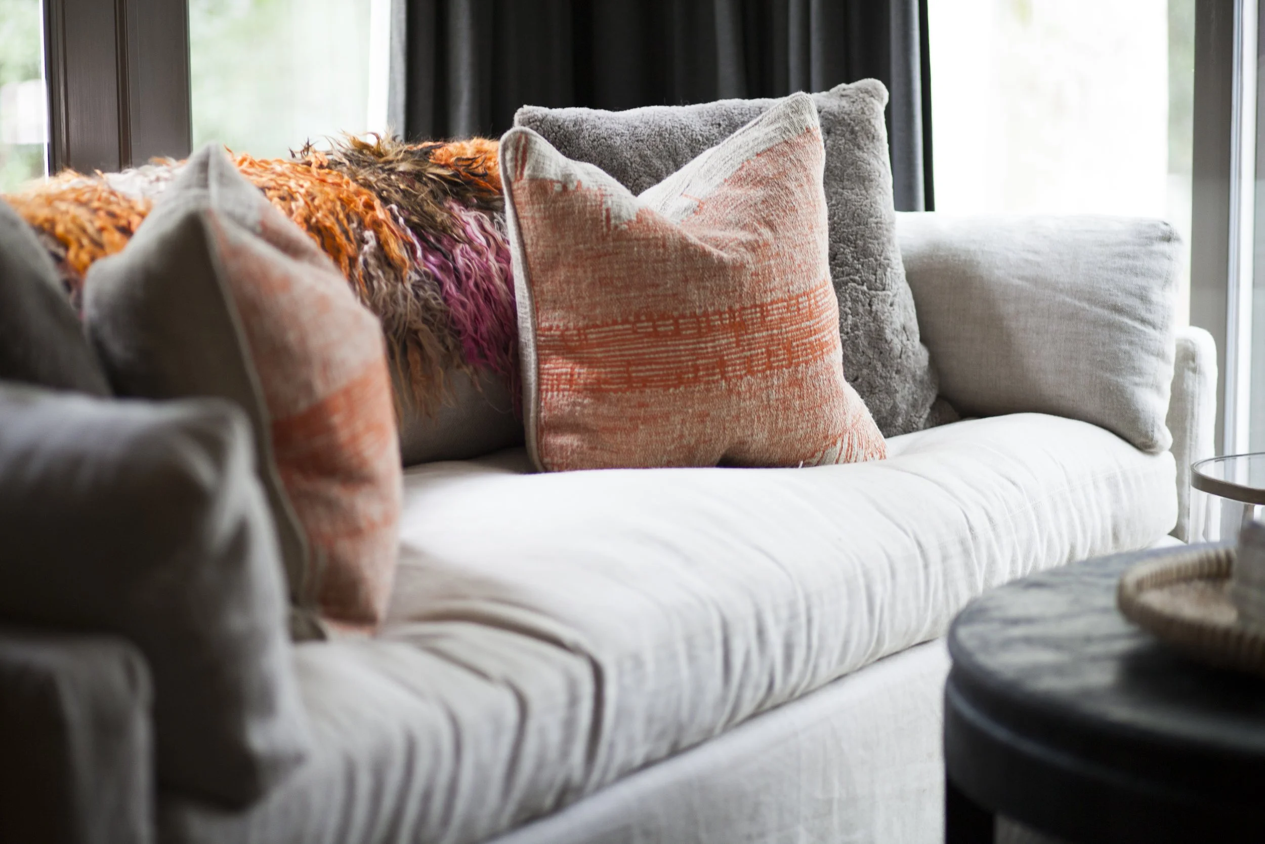 K. Interiors Design Work - A beige sofa with multiple decorative pillows in various textures and colors, with a multicolored throw blanket on top, near a window with dark curtains.