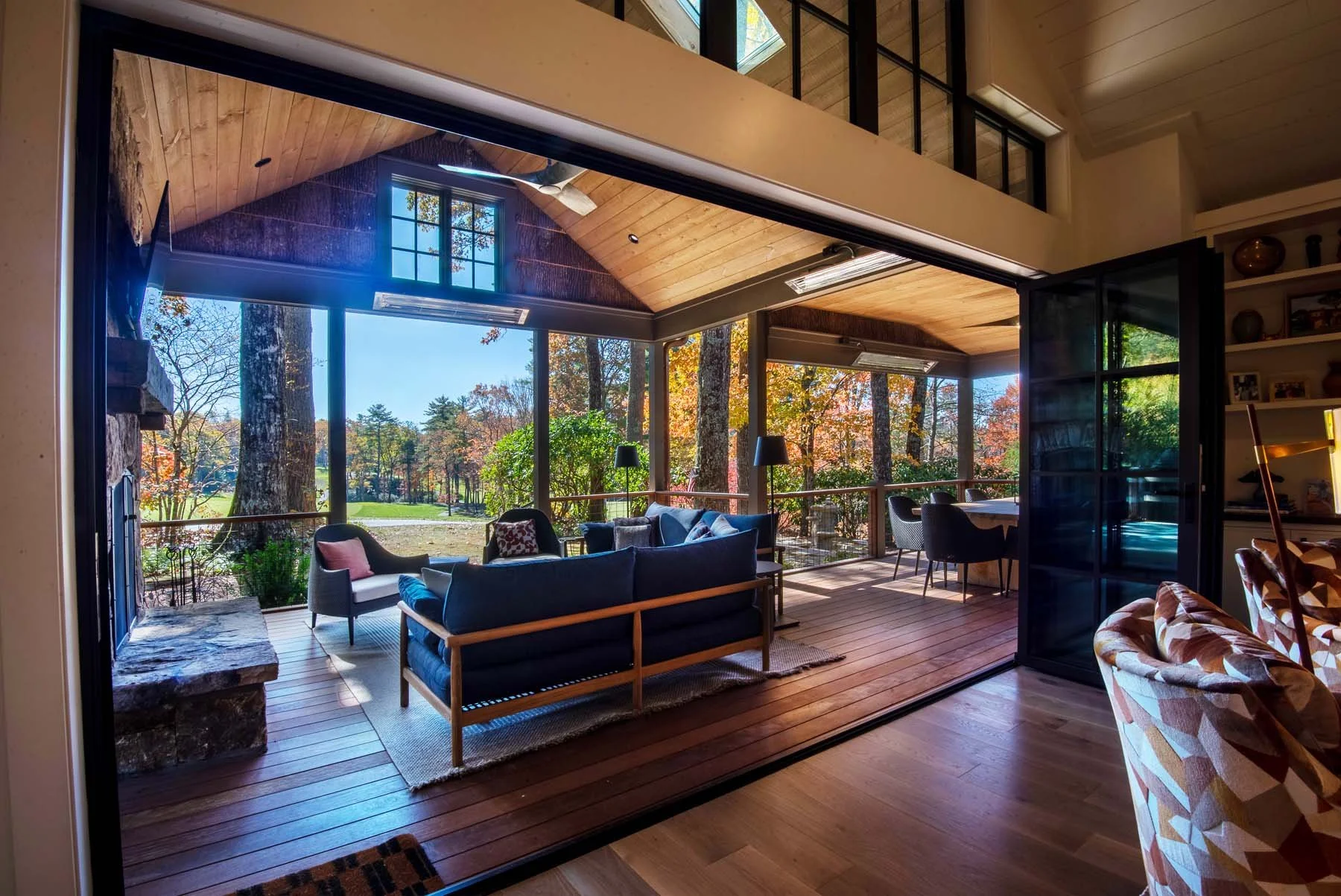 K. Interiors Design Work - Living room with open sliding glass doors leading to a screened porch with outdoor seating and view of trees with fall foliage.