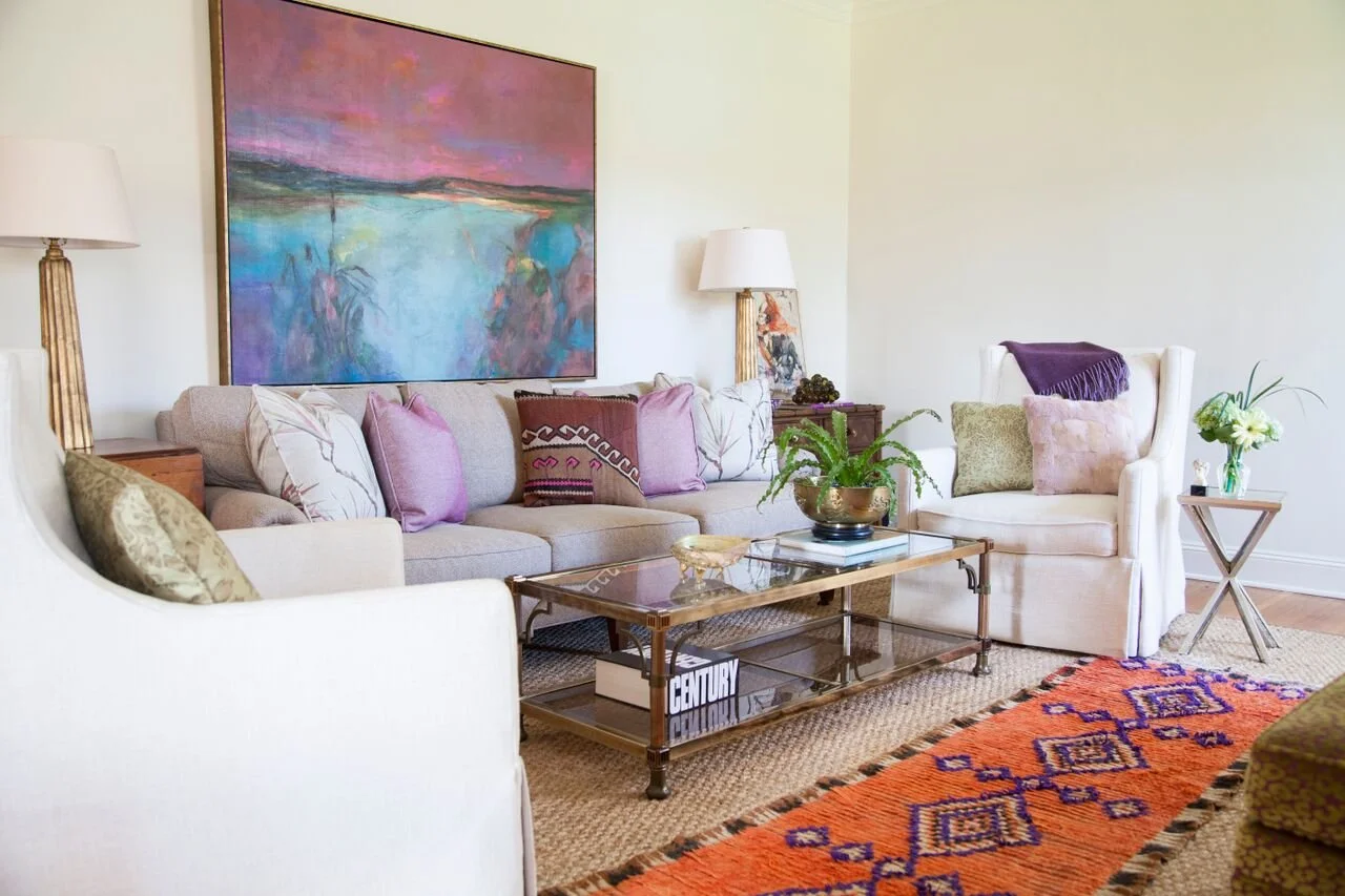 K. Interiors Design Work - Living room with beige sofa, white armchair, glass coffee table, colorful art painting, decorative pillows, side tables with lamps, potted plants, and an orange patterned rug.