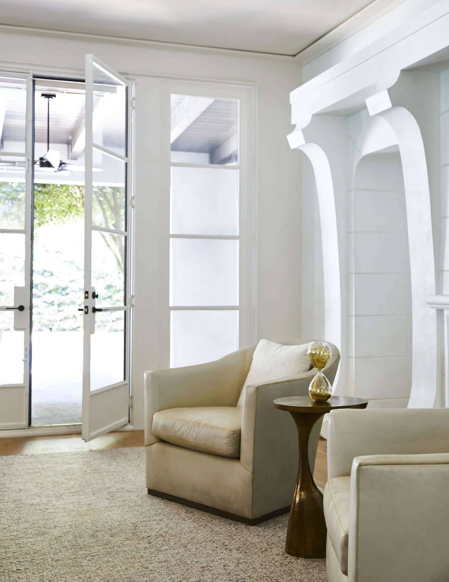 K. Interiors Design Work - Living room with open door to outside, two white armchairs, and a small wooden side table with a decorative gold hourglass.