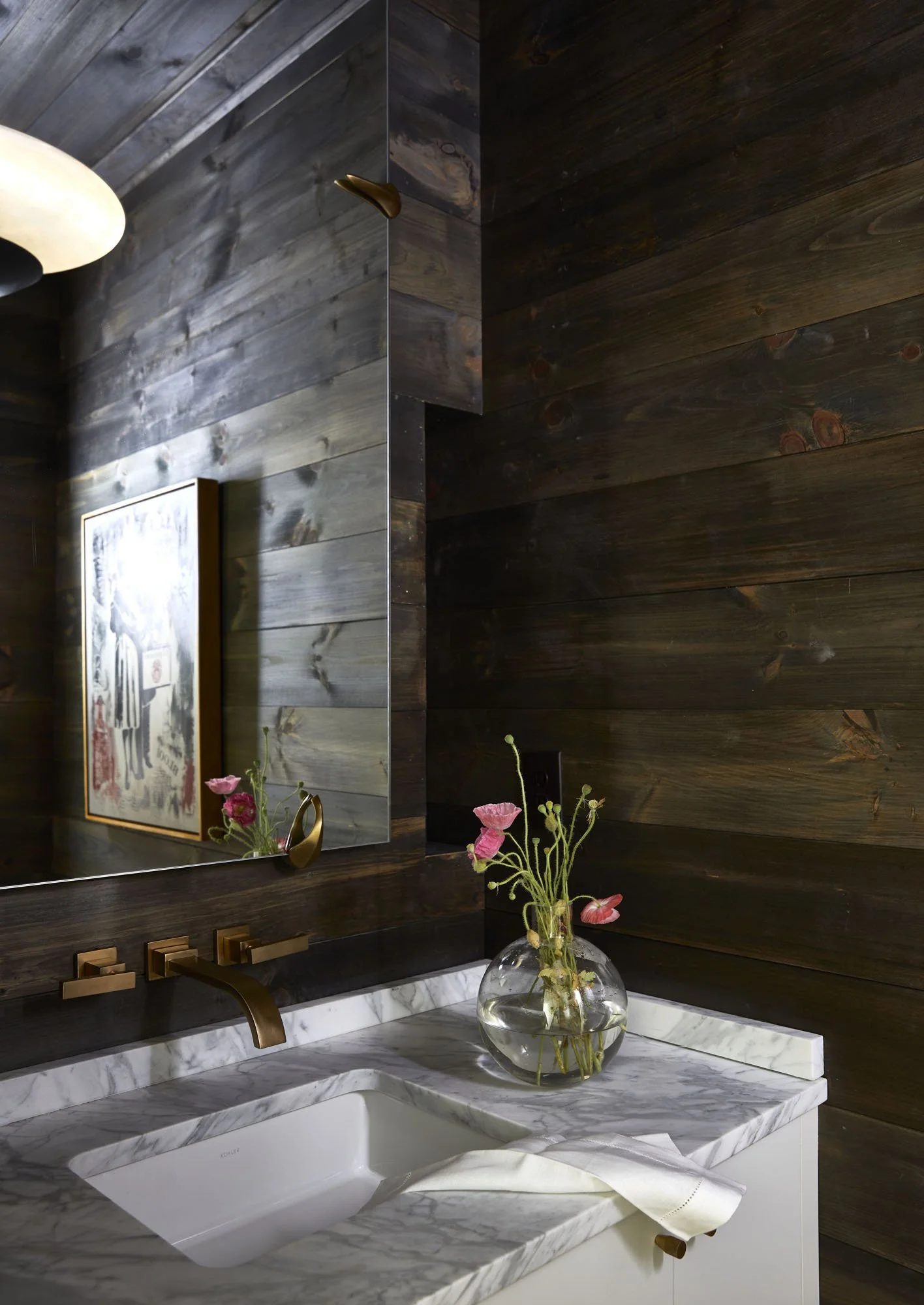 K. Interiors Design Work - Bathroom with dark wooden walls, a mirror, a marble countertop with a white sink, brass fixtures, a glass vase with pink flowers, and a framed picture on the wall.