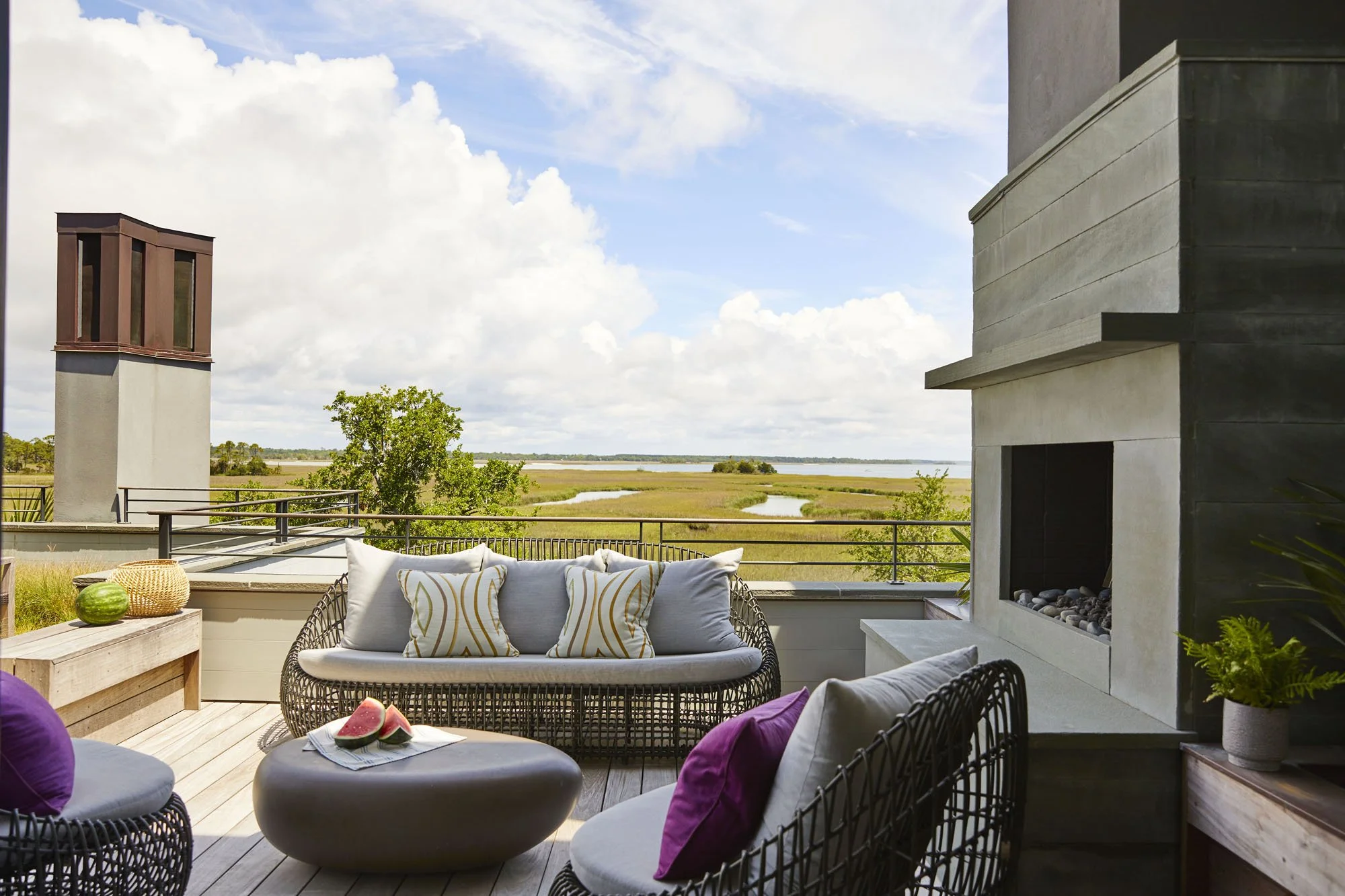 K. Interiors Design Work - Outdoor patio with wicker furniture, pillows, and a fireplace overlooking marshland and water under a cloudy sky.