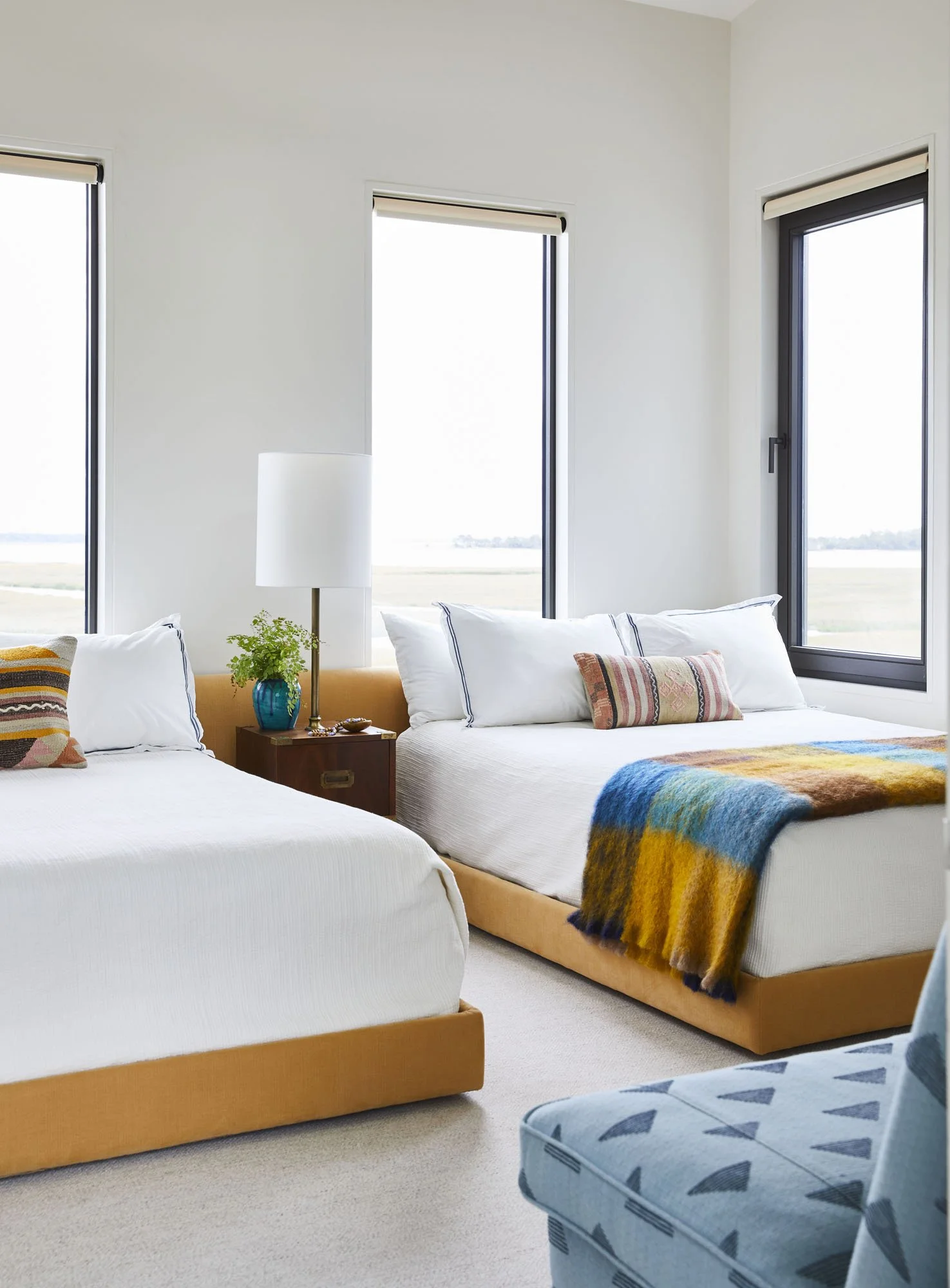 K. Interiors Design Work - A bright bedroom with two beds, a nightstand with a blue vase and lamp, and large windows showing an outdoor landscape.