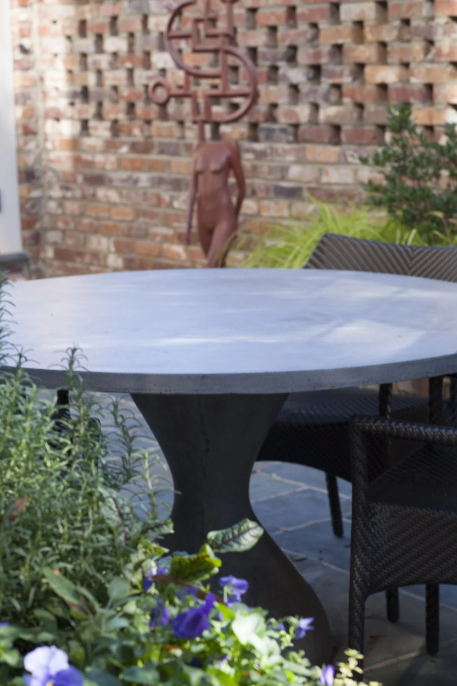 K. Interiors Design Work - Outdoor patio table with wicker chairs, surrounded by green plants and purple flowers, brick wall with metal sculpture in the background.