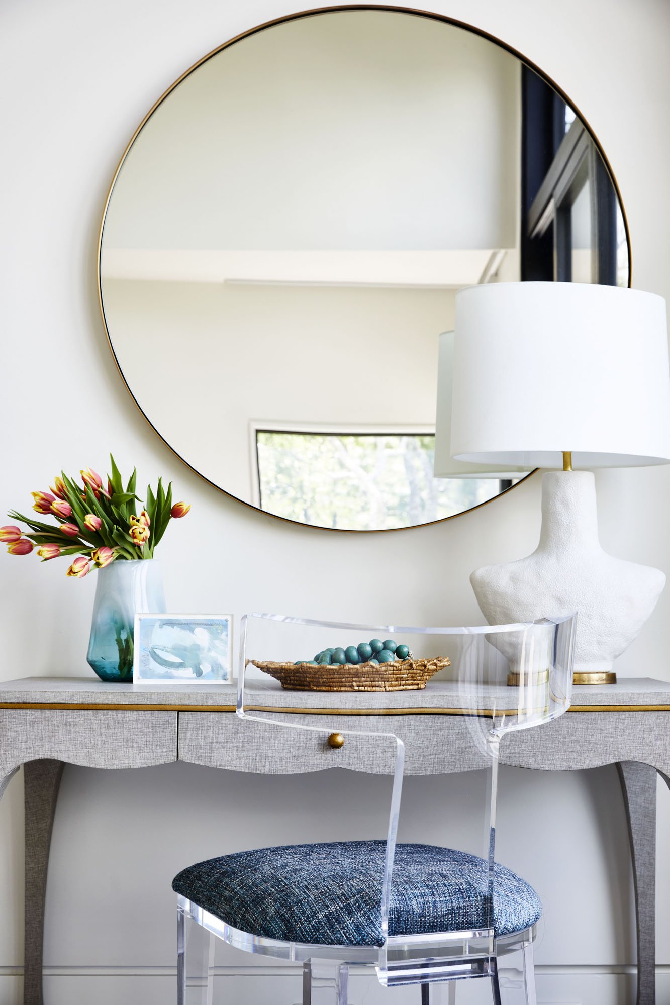 K. Interiors Design Work - A contemporary living room console table with a large round mirror above, a white table lamp, a vase of pink tulips, a framed photo, and a decorative beaded bowl, reflecting a window and greenery outside.