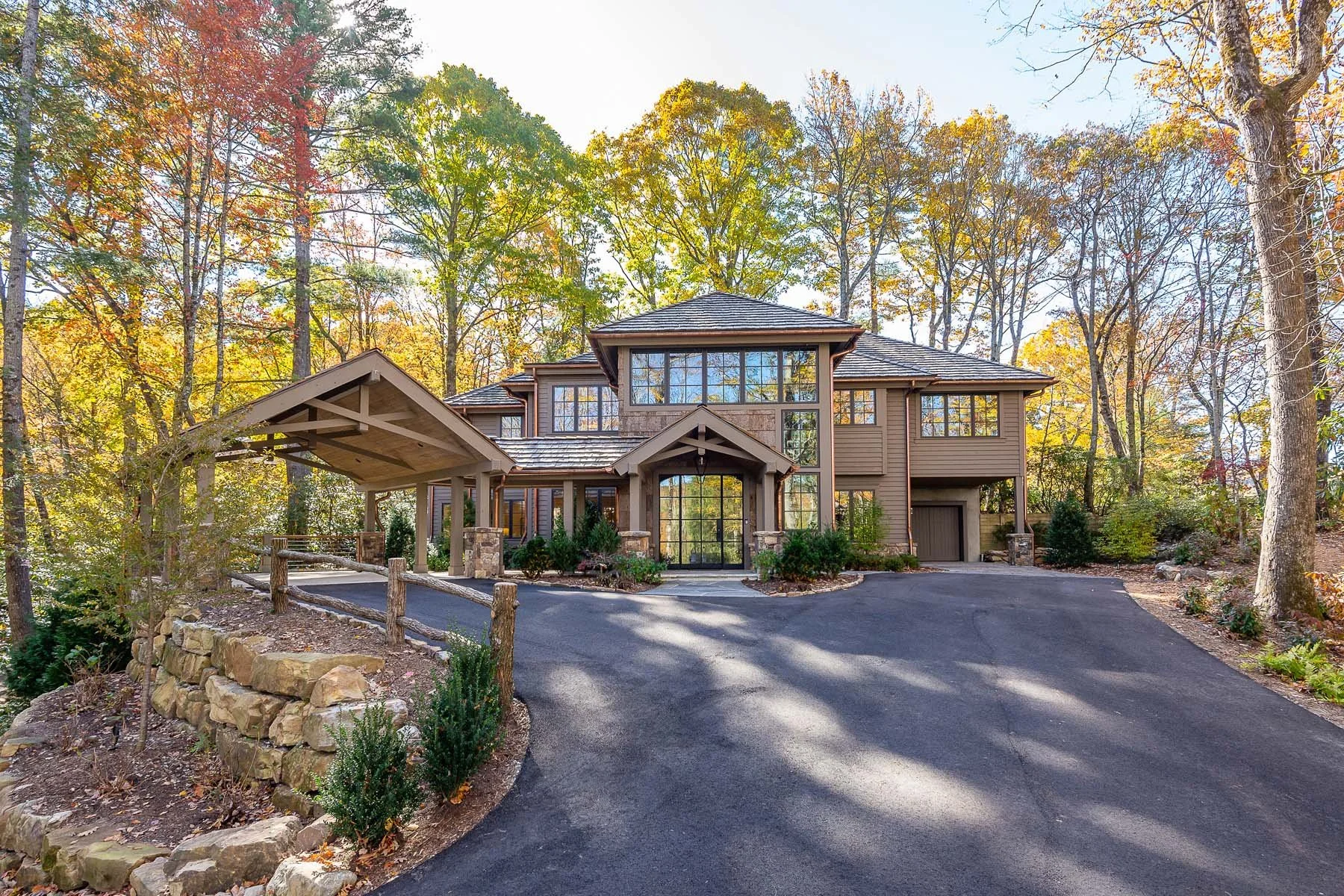 K. Interiors Design Work - Large modern house with stone and wood exterior, multiple windows, and a peaked roof, set in a wooded area with tall trees and a paved driveway.