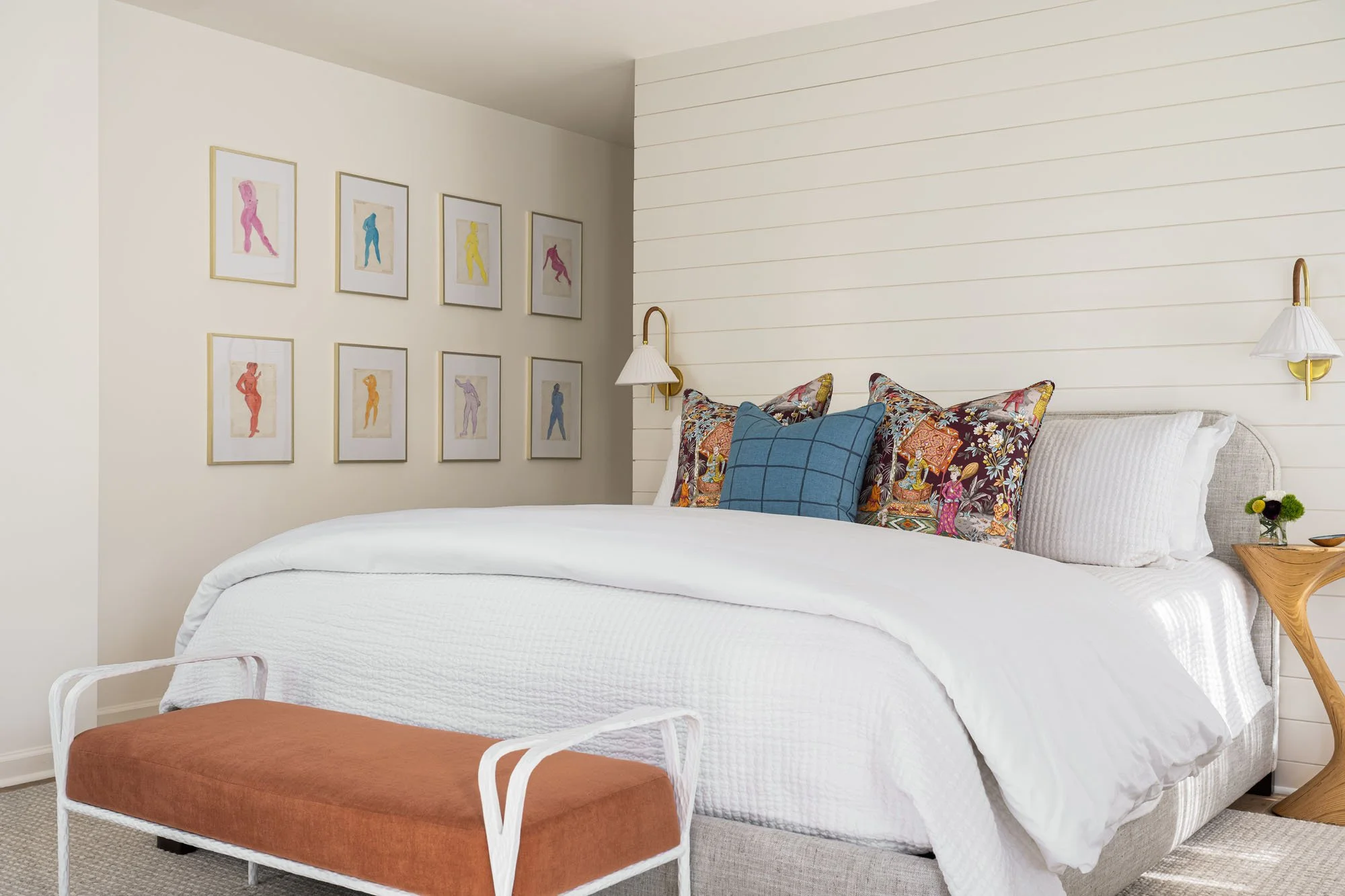 K. Interiors Design Work - A neatly made bed with white bedding, decorative pillows, and a wooden bench at the foot of the bed in a modern bedroom with artwork on the wall.