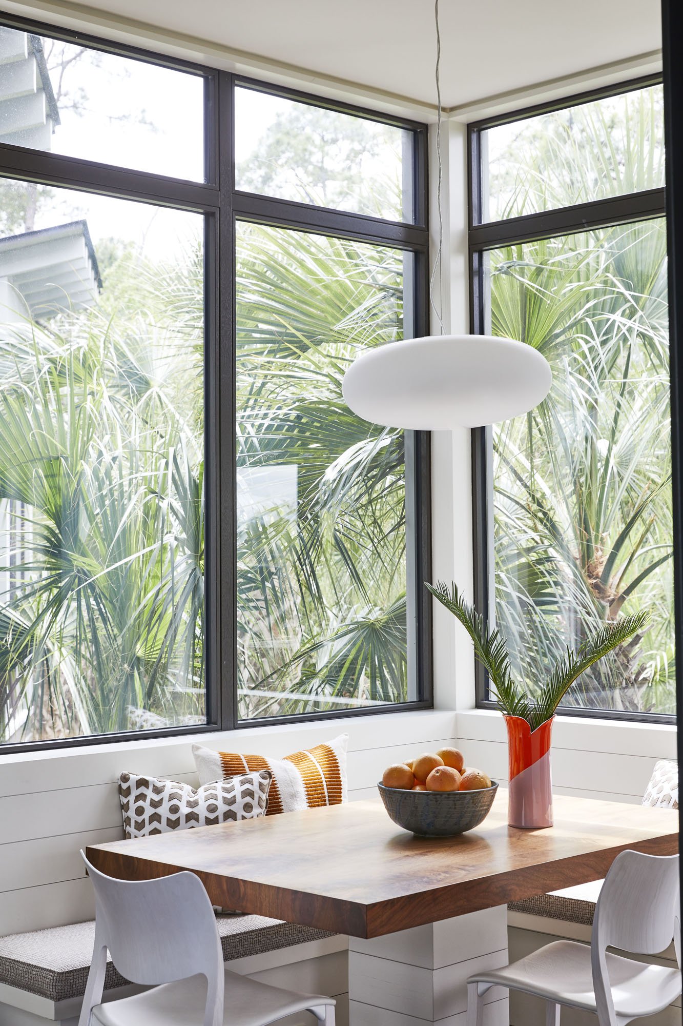 K. Interiors Design Work - Bright dining nook with a wooden table, white chairs, decorative pillows, a ceramic bowl of oranges, a red and pink vase with green palm leaves, and large corner windows with views of lush tropical plants outside.
