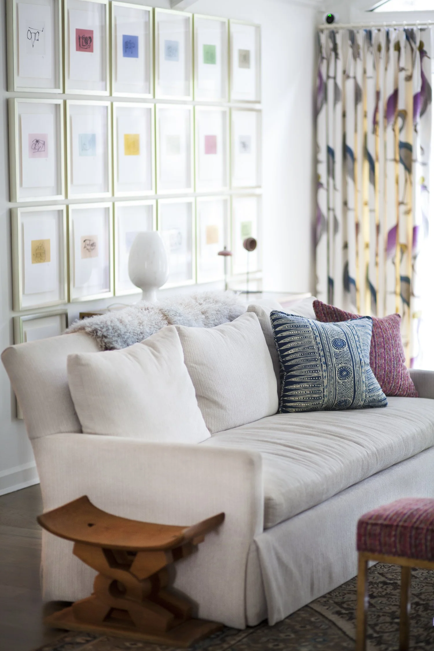 K. Interiors Design Work - Living room with a beige sofa decorated with colorful pillows, a wall with framed artwork, a wood side table, and a floral curtain.